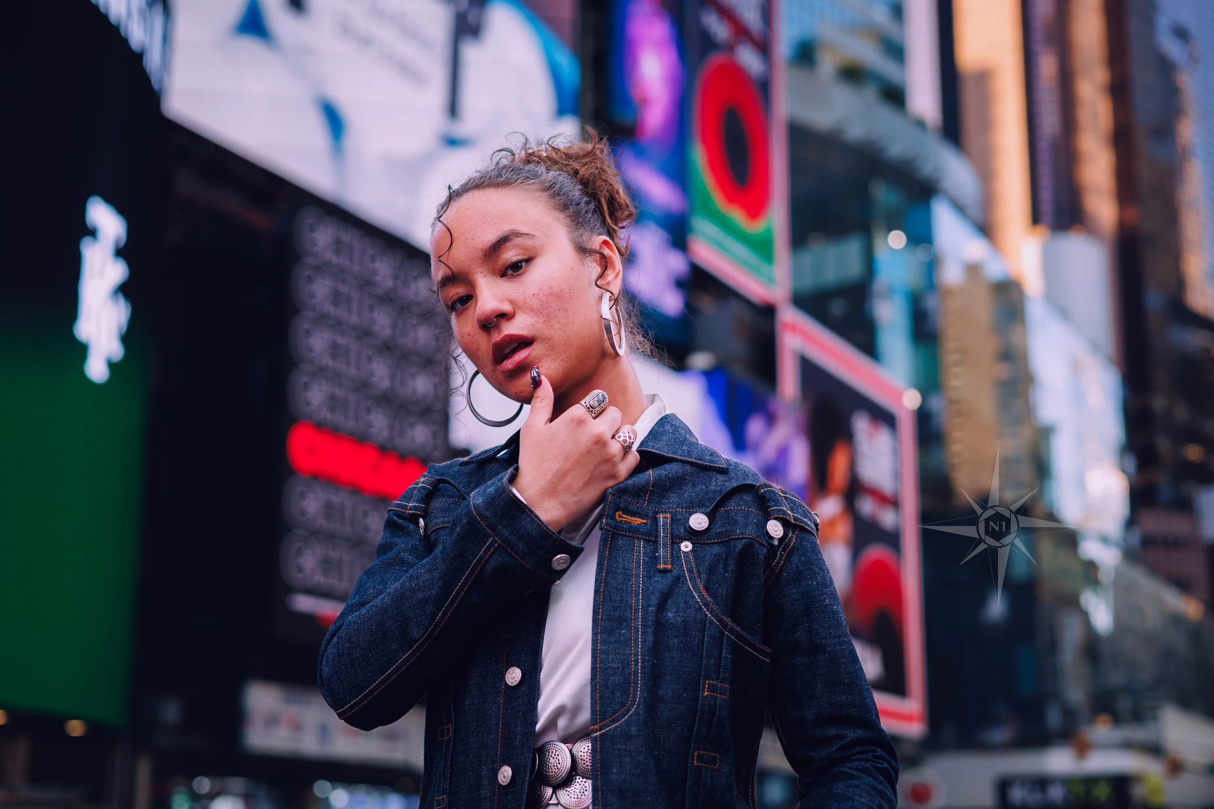 A young woman with curly hair and large hoop earrings standing in Times Square, New York City, surrounded by bright electronic billboards and city buildings.