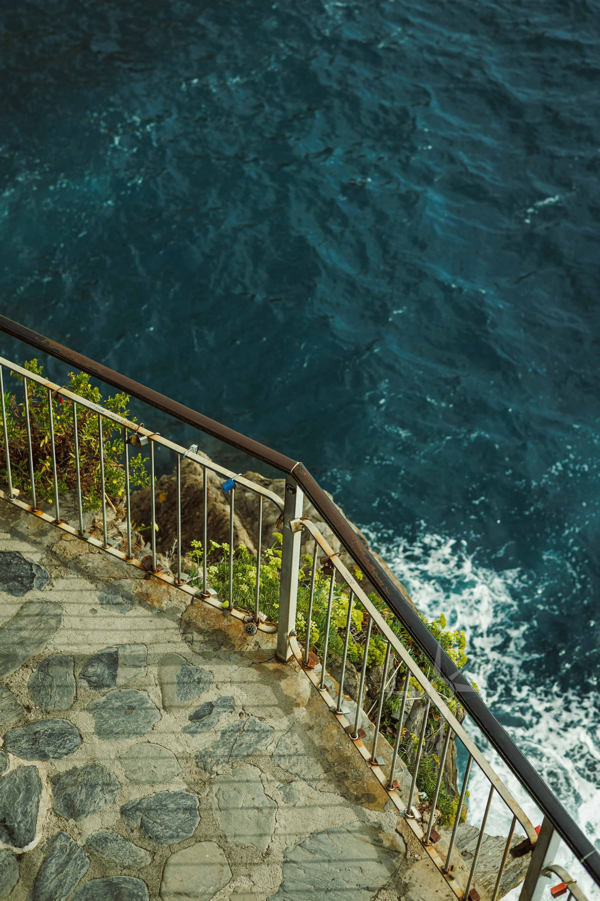 View of a rocky stone walkway with a metal railing overlooking the ocean, with the water an intense blue and gentle waves.