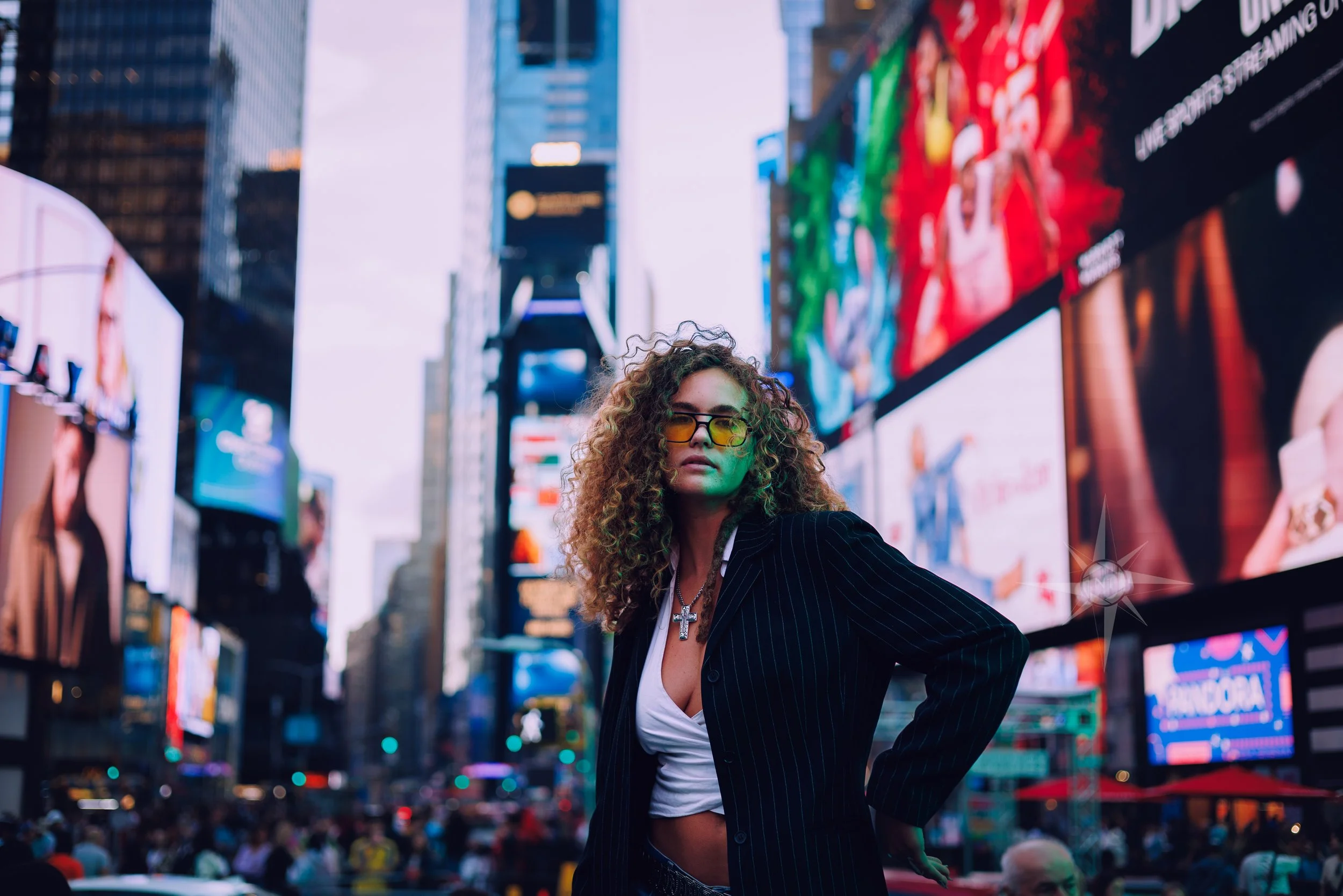 A woman with curly hair wearing sunglasses, a white crop top, and a black pinstripe blazer stands in Times Square, New York City, surrounded by lit-up billboards and electronic advertisements.