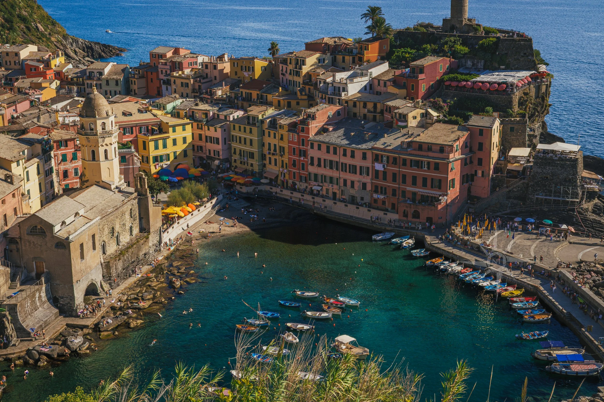 Colorful seaside village with boats docked in clear blue water, pastel buildings, a church with a bell tower, and a small beach area with people.