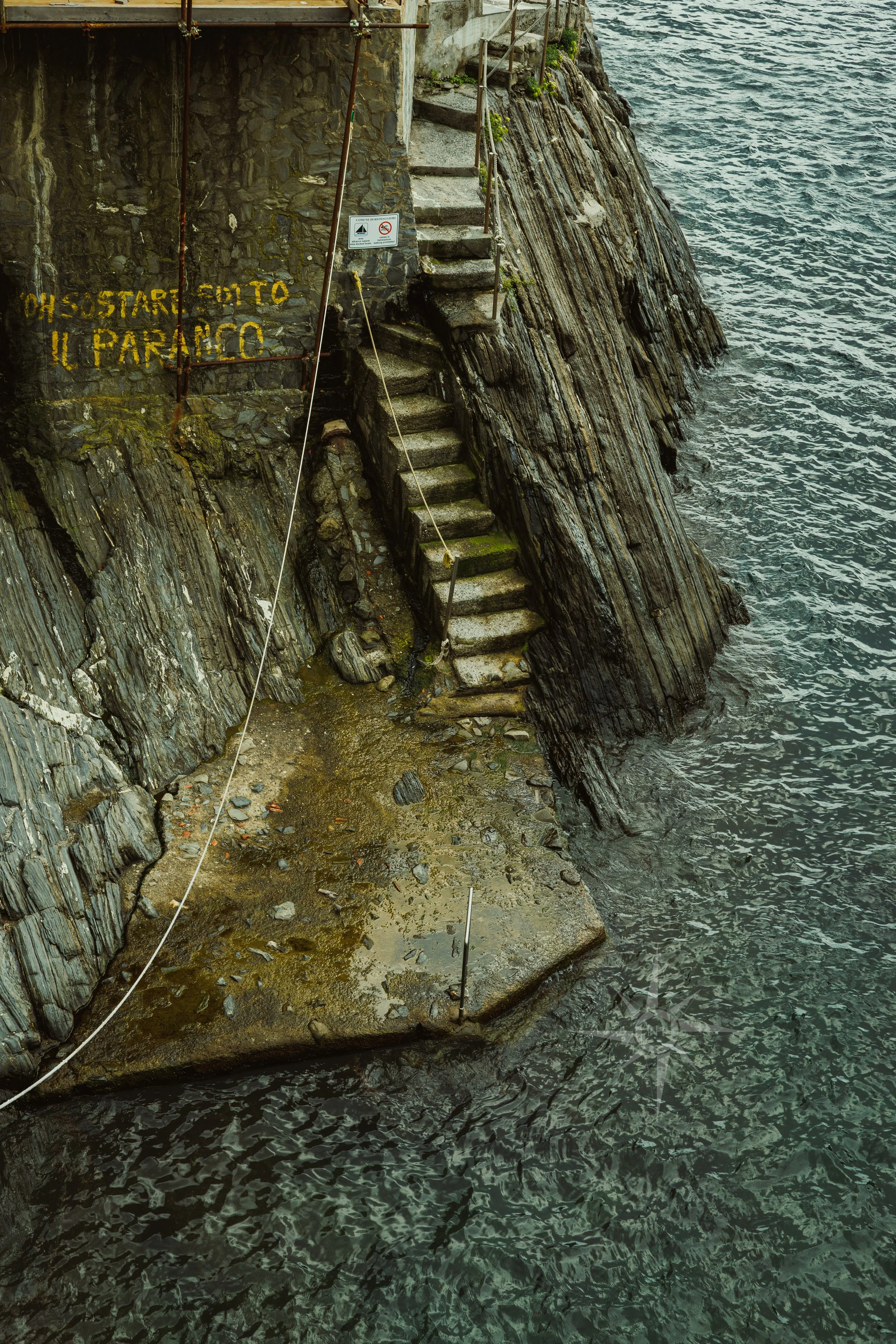 Concrete stairs leading up a rocky cliffside by the water, with caution signs and a yellow-painted message in Italian on the cliff.