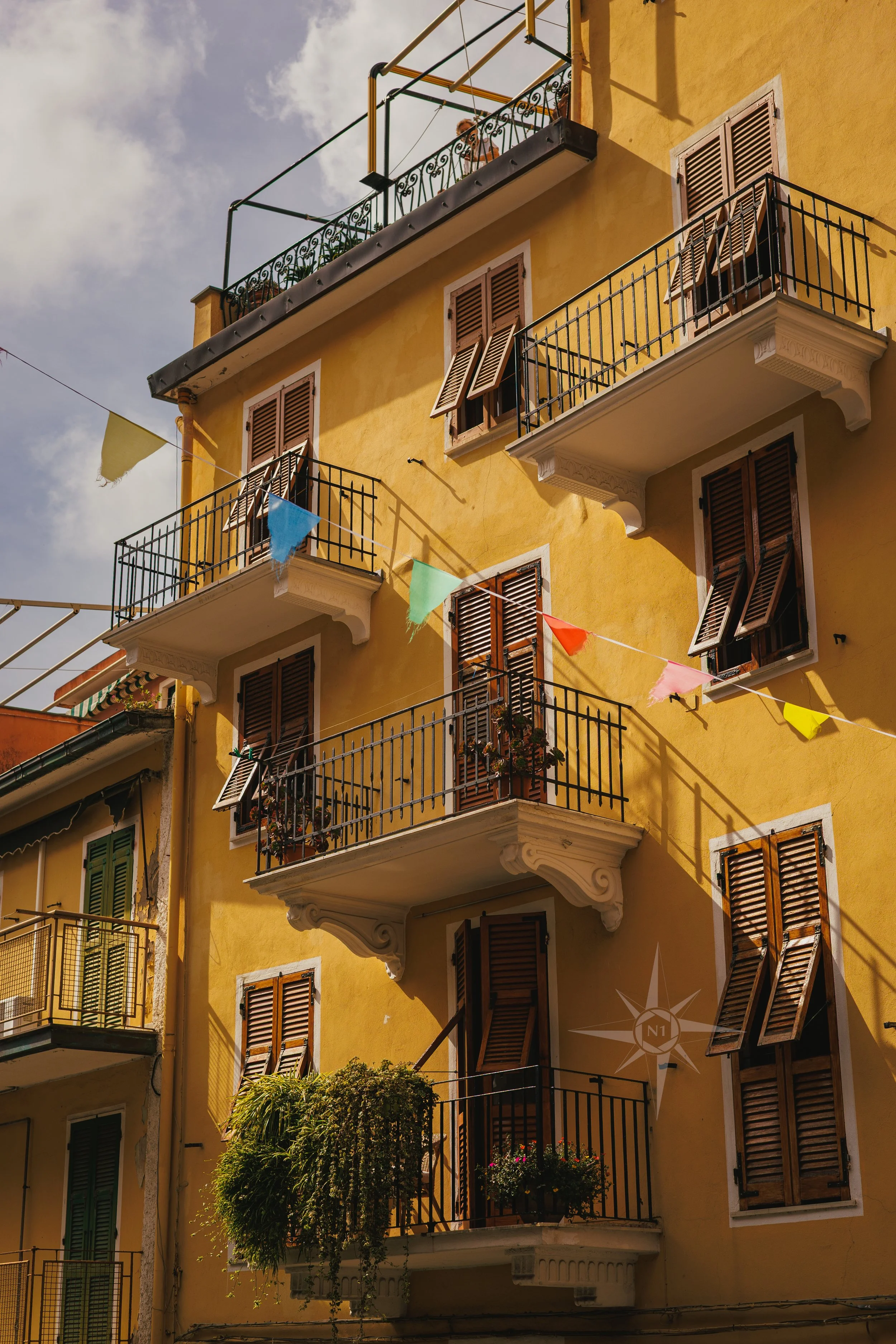 A yellow building with brown shutters and several balconies, decorative iron railings, and colorful bunting flags strung across the facade.