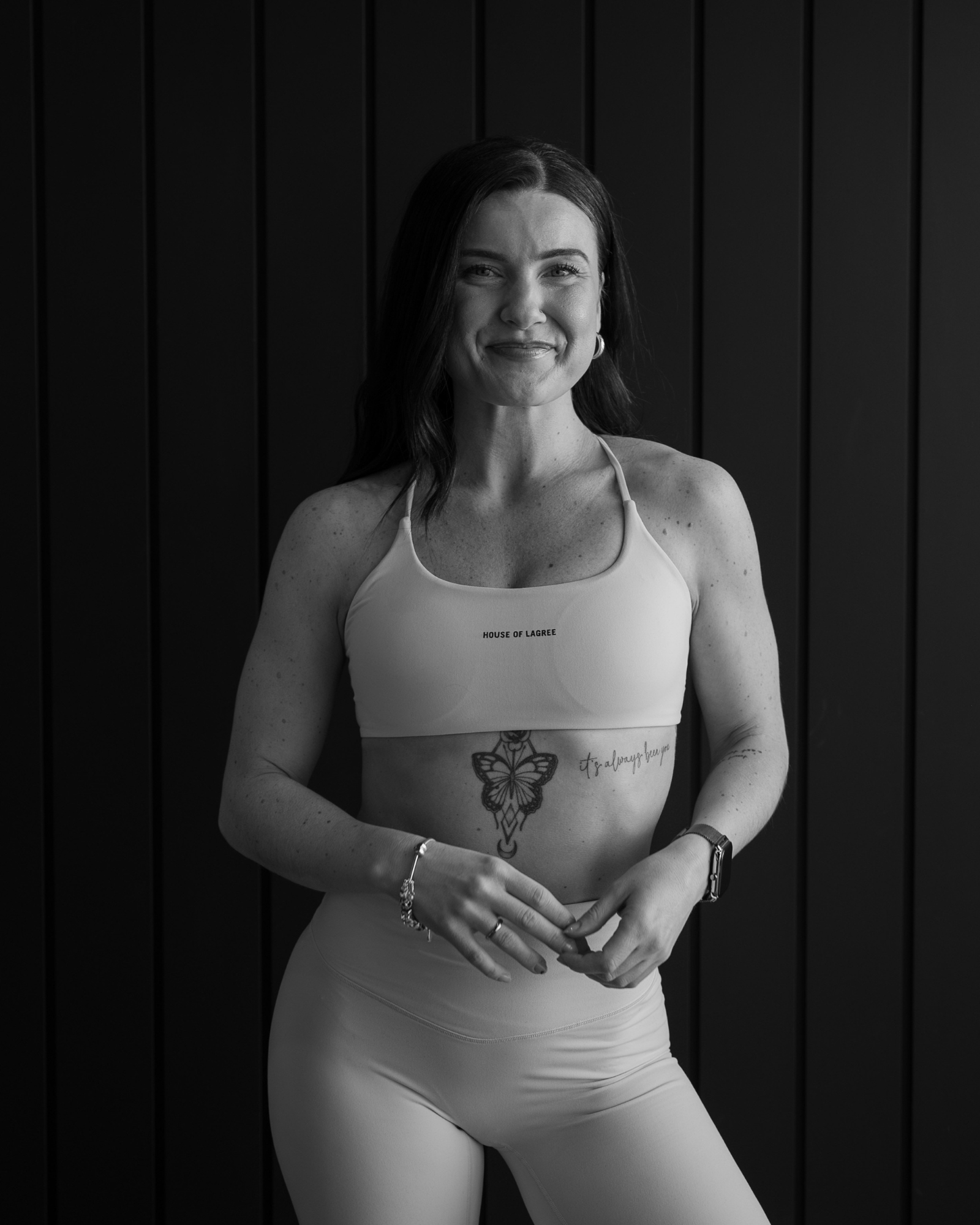 Black and white photo of a smiling woman with long dark hair, wearing a sports bra and leggings, standing in front of a dark wooden wall.