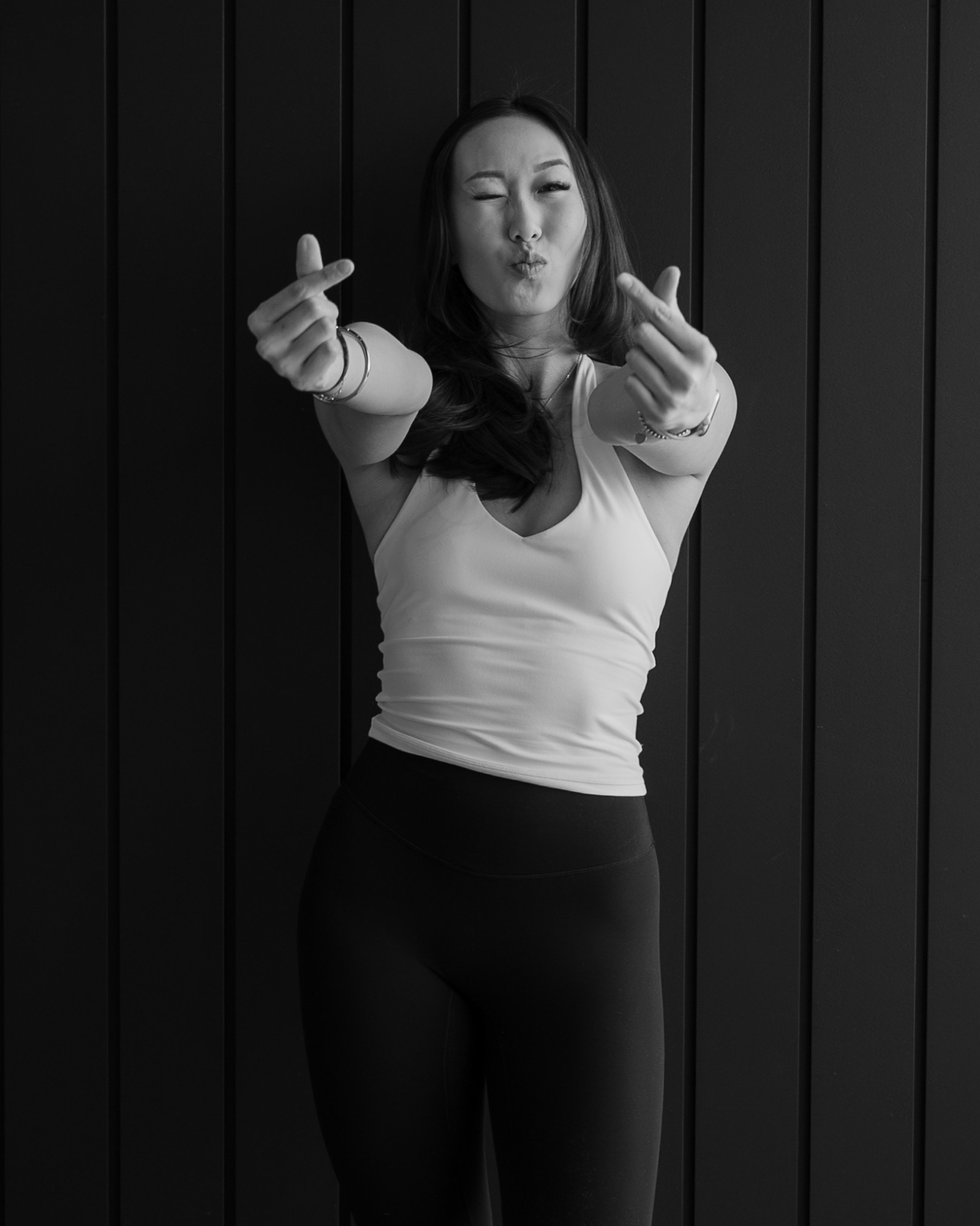 A woman in a white tank top and black pants making a playful kissing face and heart gestures with her hands, standing against a dark wooden wall.