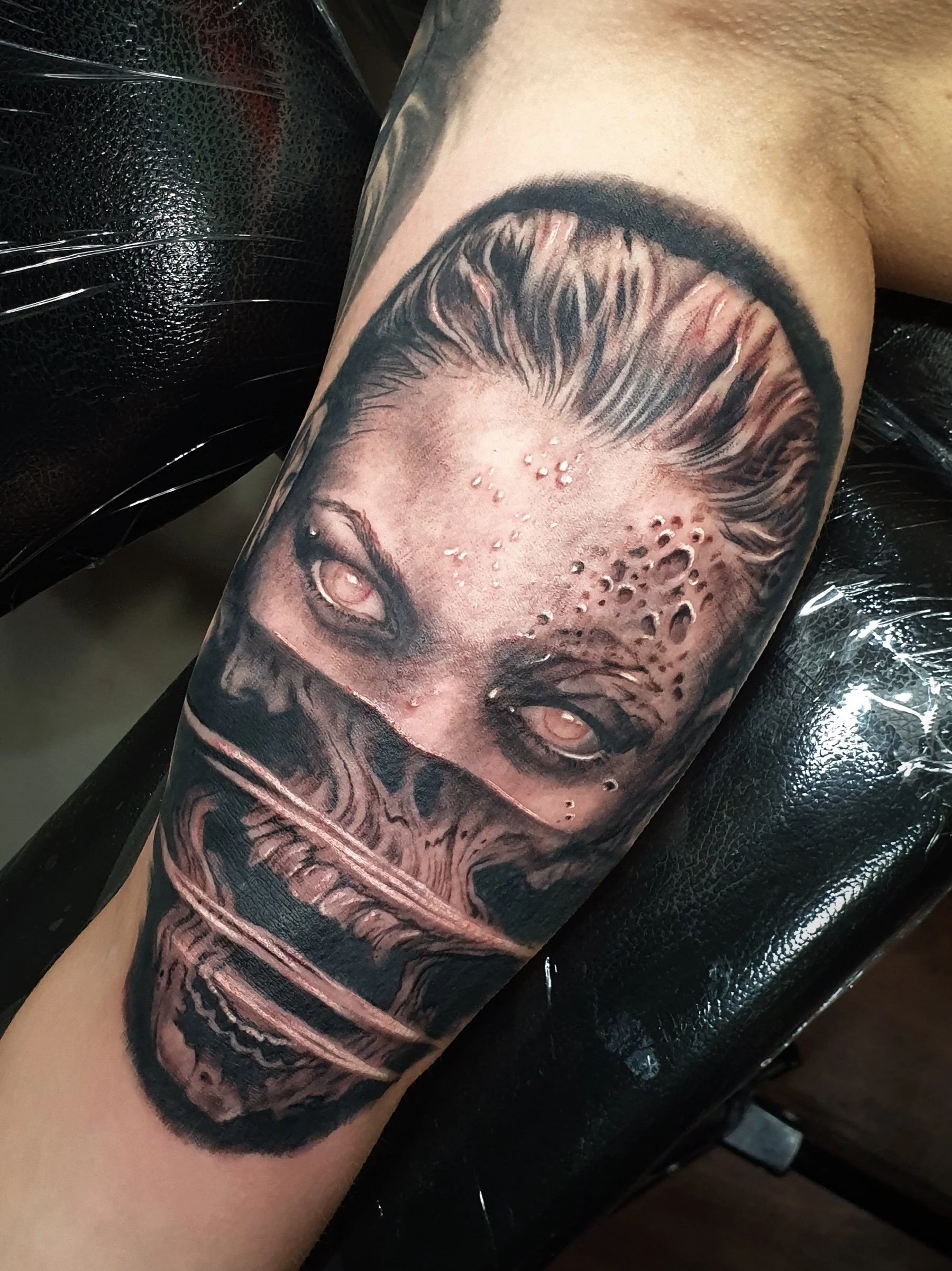 Black and grey tattoo of a female face with piercing eyes, hair styled up, and a face partly covered by a hand, on a person's arm.