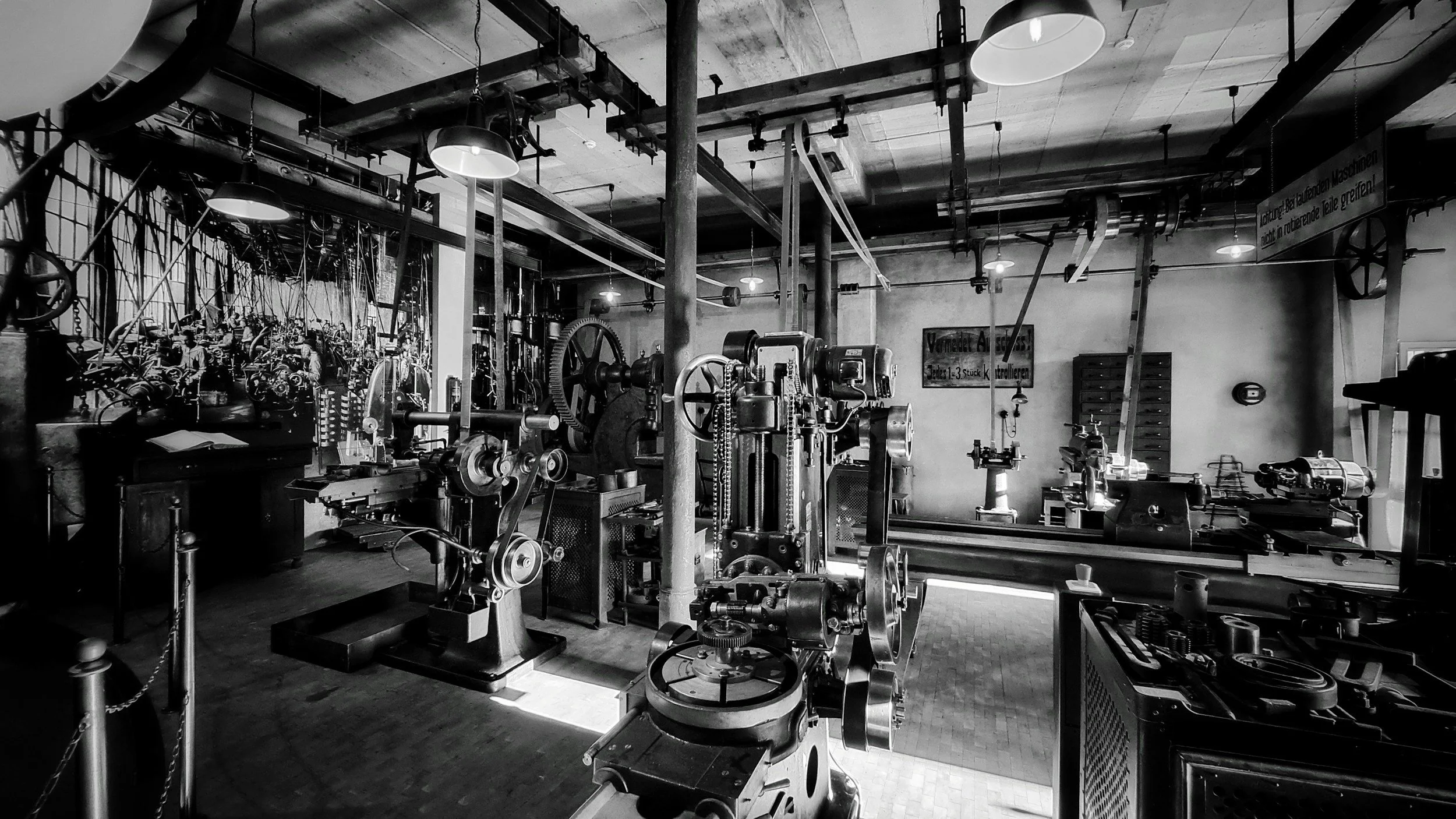 Black-and-white view of a historic industrial machine shop filled with vintage lathes, milling machines, gears, pipes, and overhead belt-driven machinery.