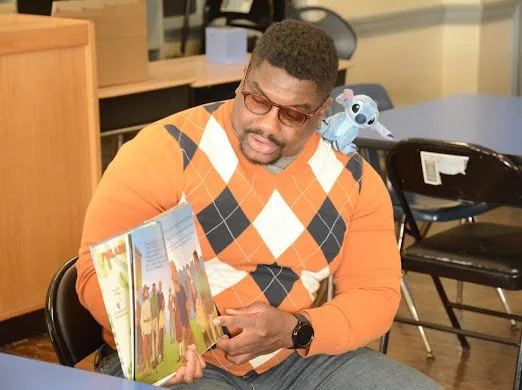 A man with glasses and a ruffled beard reading a children's book at a table, with a Stitch plush toy on his shoulder.