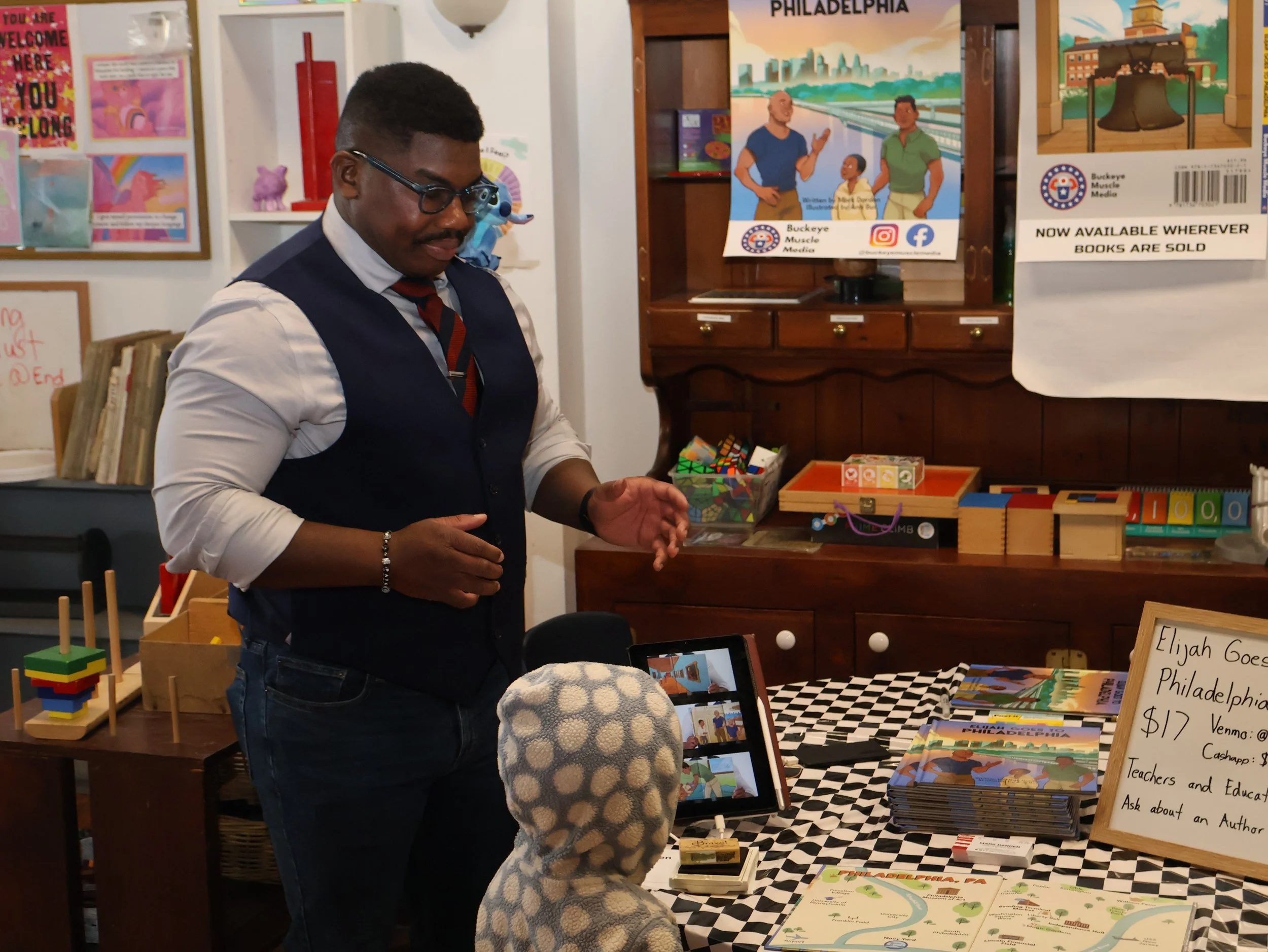 A man in a business vest and glasses is standing at a table with books and educational materials, speaking to children at a corner of a bookstore or event space with posters and signs on the wall, including one for "Elijah Goes to Philadelphia."