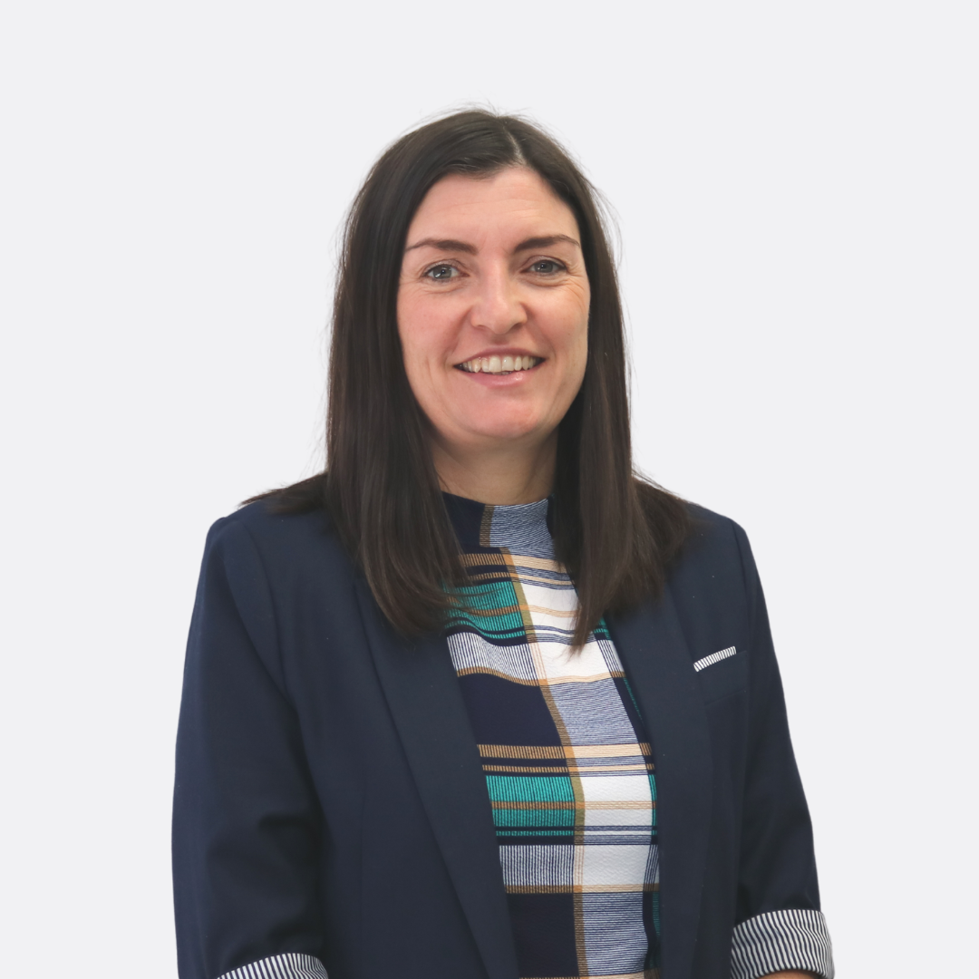 A woman with shoulder-length dark hair, wearing a dark blazer and a multicolored plaid shirt, smiling in front of a plain white background.