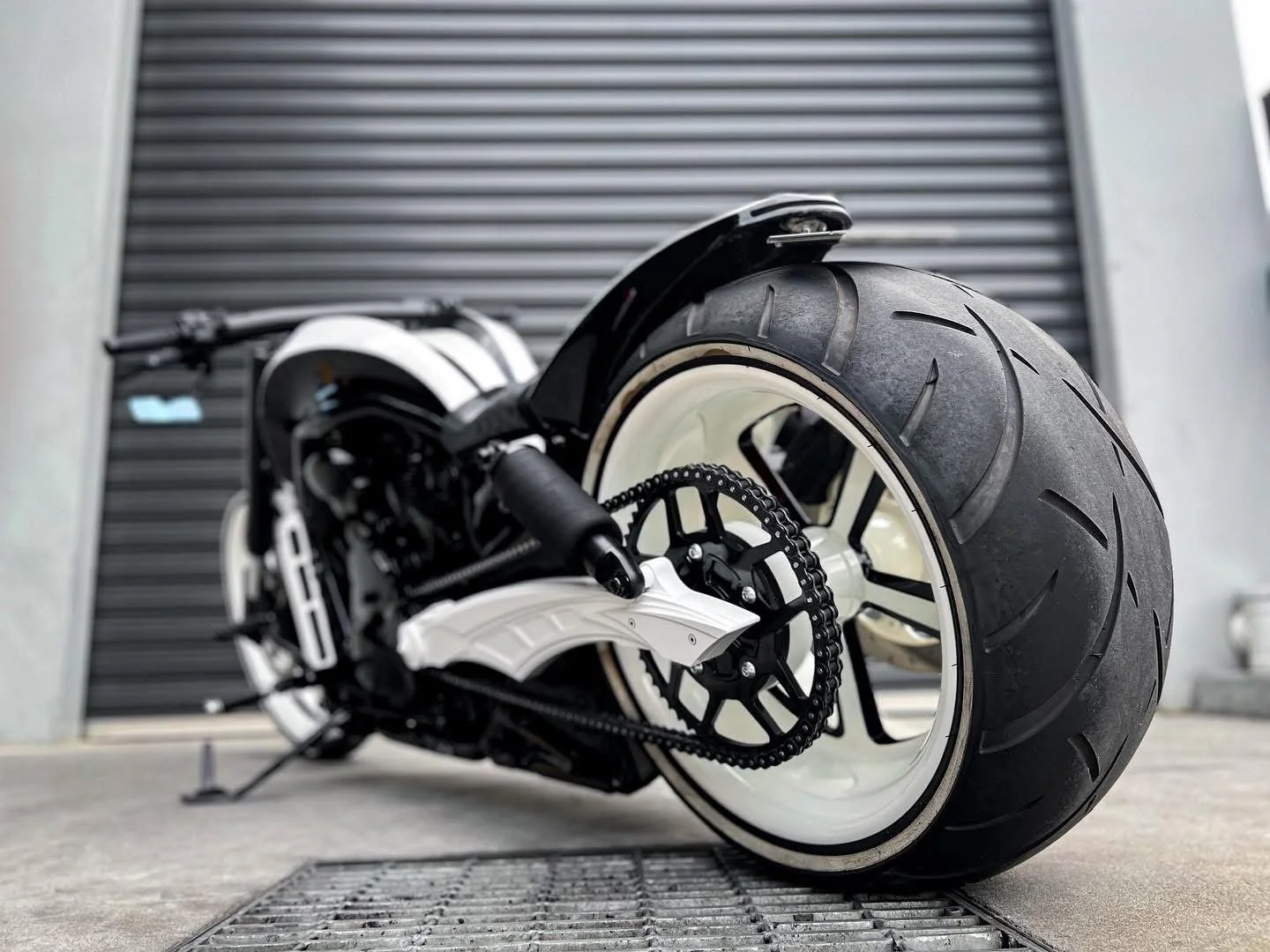 Close-up of a modern motorcycle with wide rear tire and black and white color scheme, parked on a concrete surface with a metal grate and roller shutter in the background.