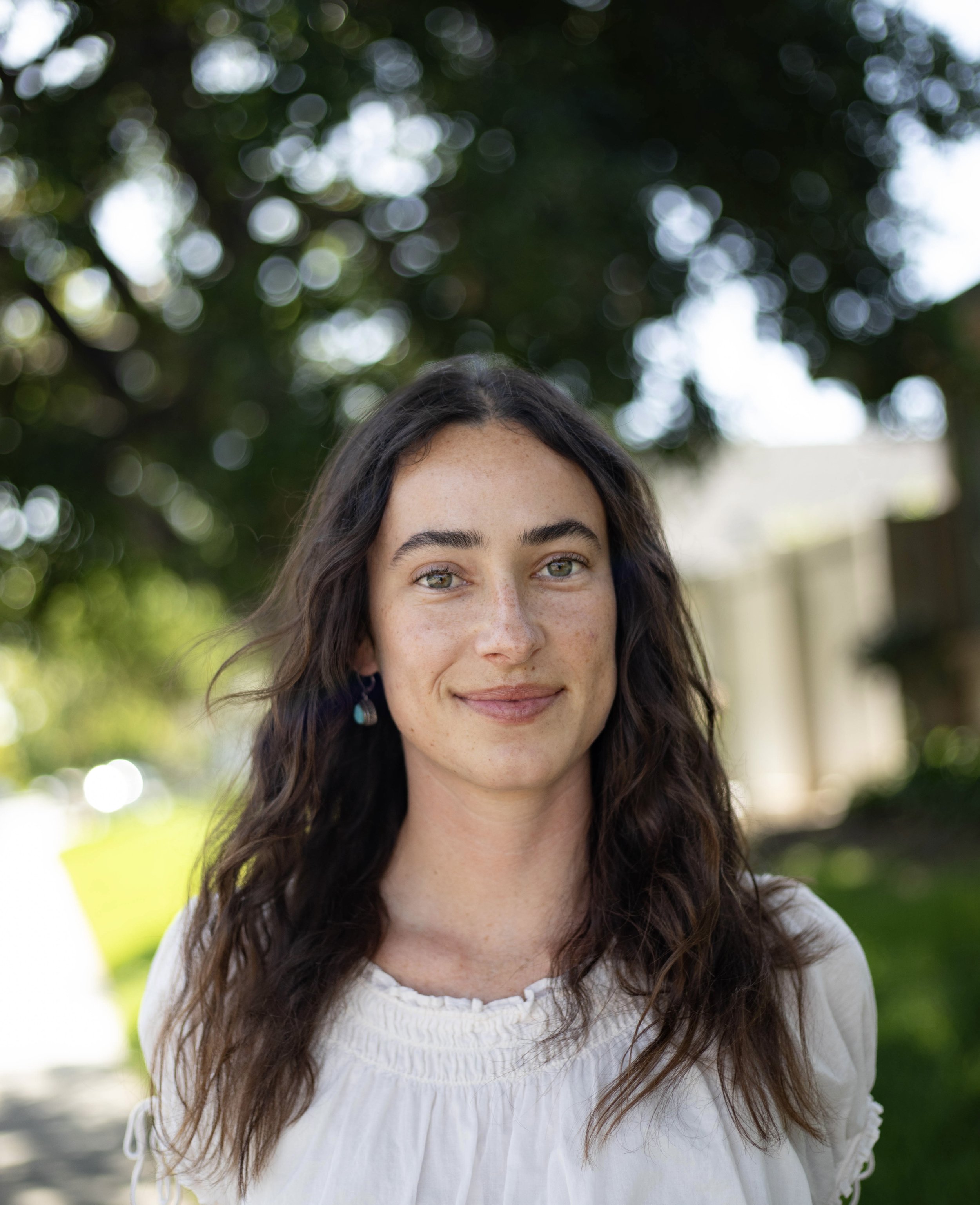 A woman with long brown hair and bangs, smiling, sitting in front of a large green leafy plant, wearing a checkered shirt and light blue jeans.