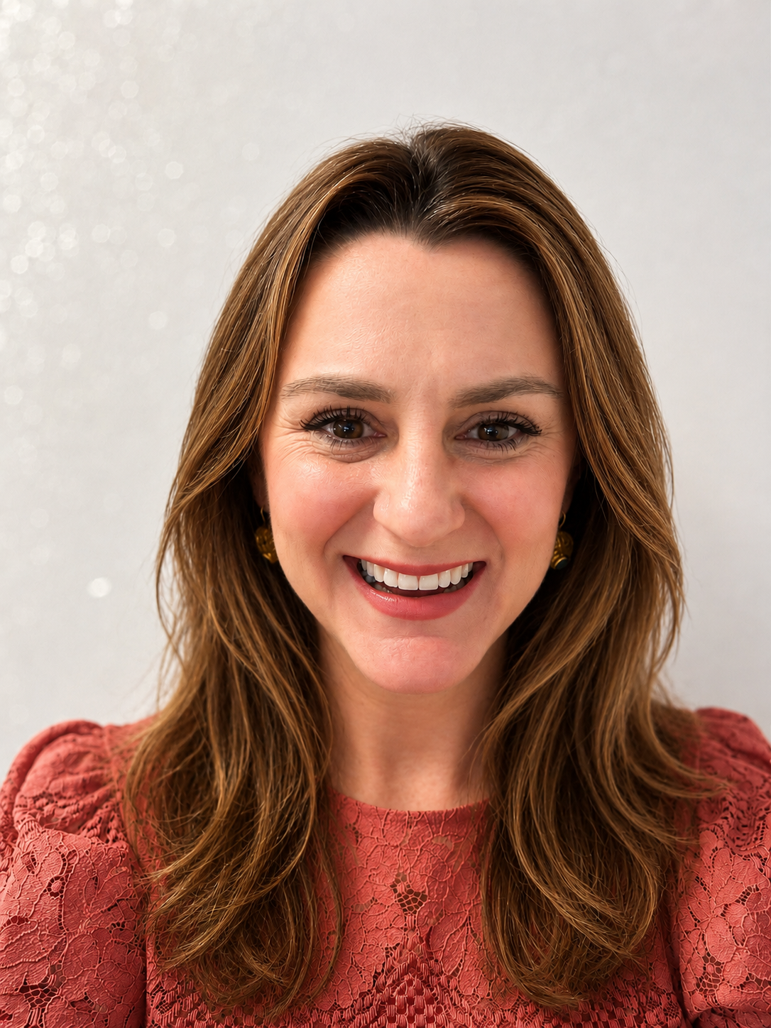 Close-up of a smiling woman with brown hair, light makeup, and earrings, outdoors with blurred background.