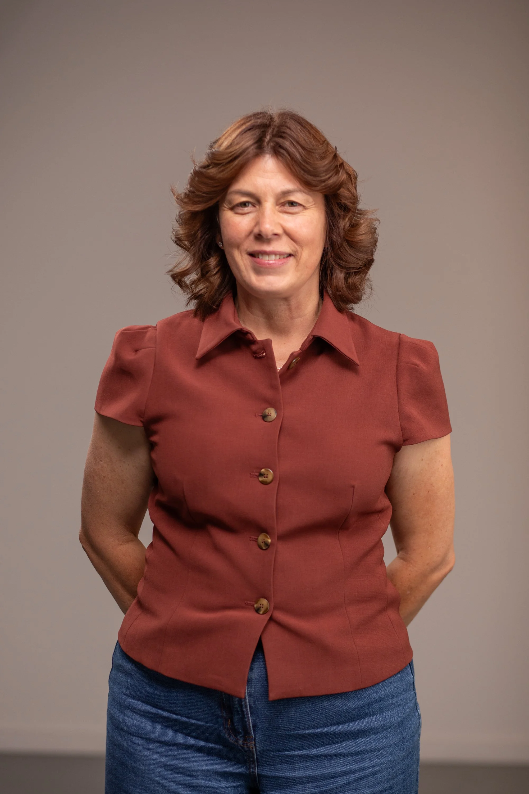 A woman with short curly brown hair smiling, wearing a sage green dress with ruffled sleeves and a long beaded necklace, standing against a plain white background.
