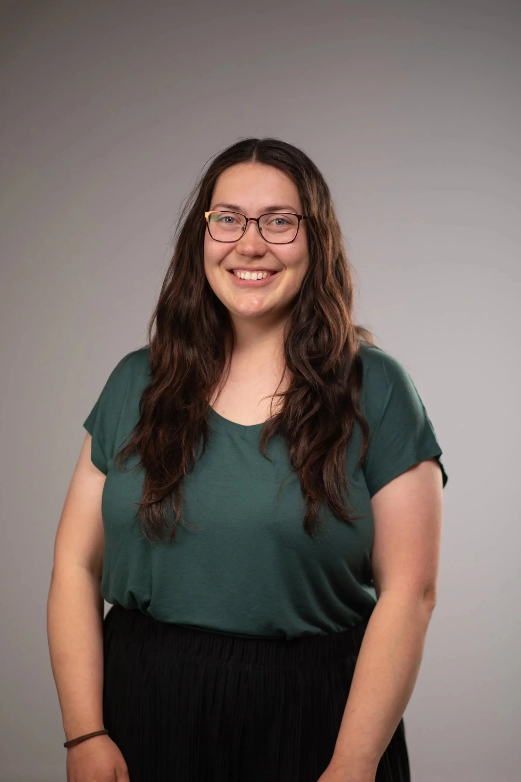 A smiling woman with long wavy brown hair, wearing glasses and an orange button-up shirt, stands against a plain light gray background.