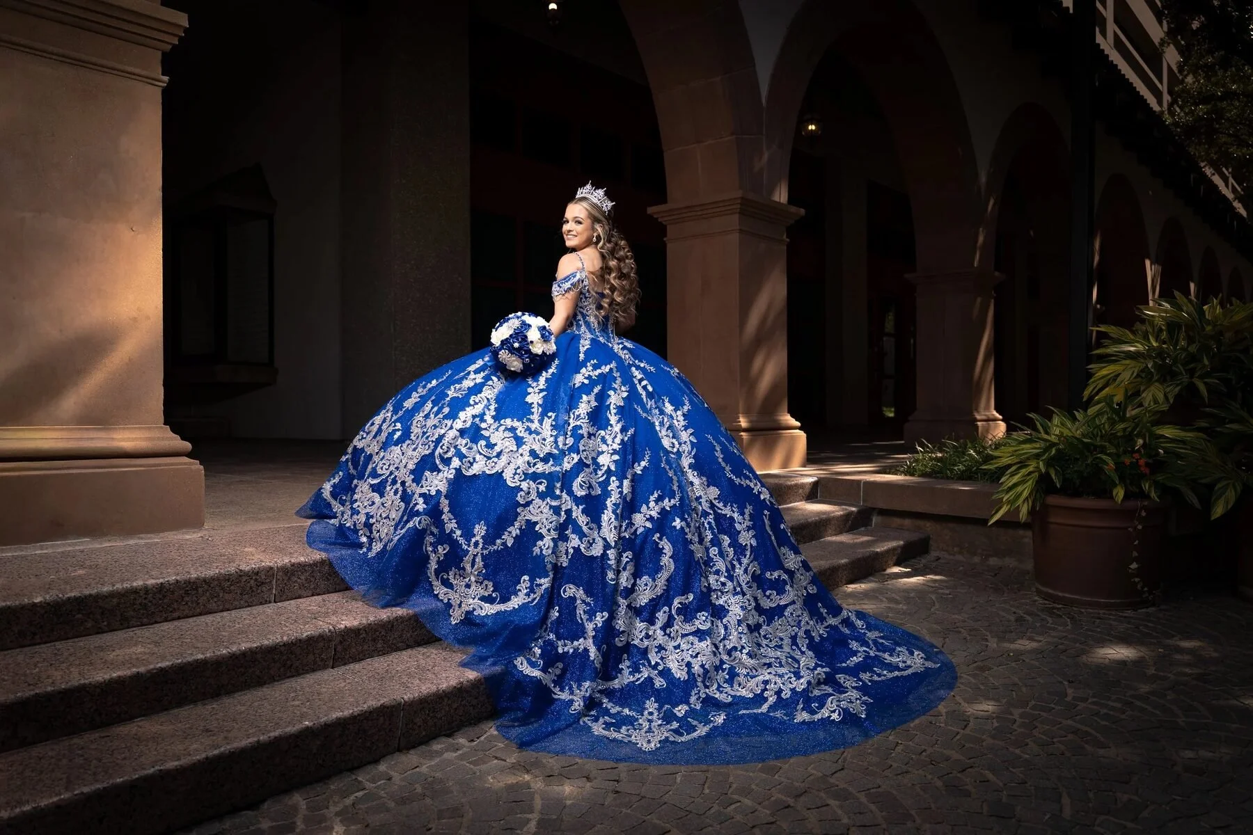 Quinceañera in a royal blue and silver gown on steps at a Dallas villa