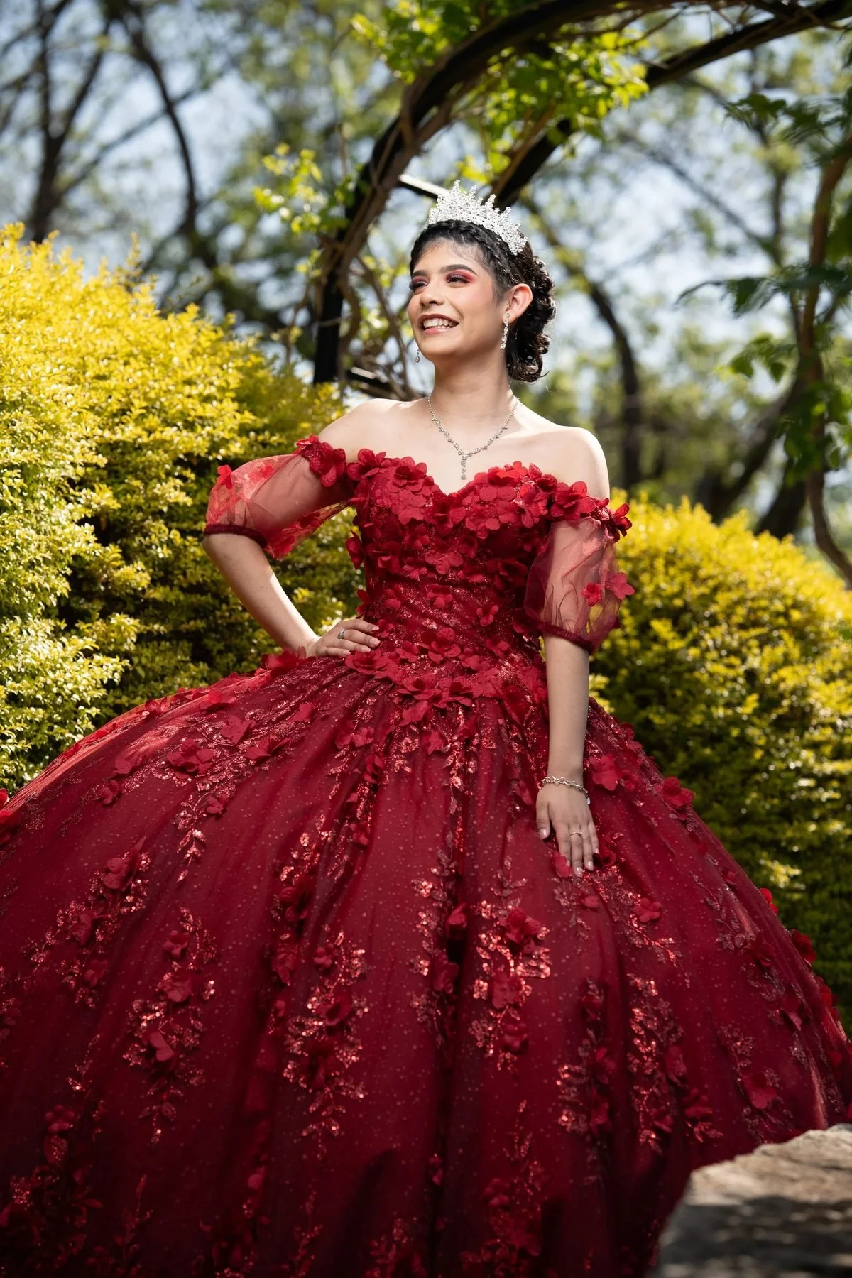Quinceañera in a red 3D floral ball gown and tiara standing in a sunlit garden in Texas.