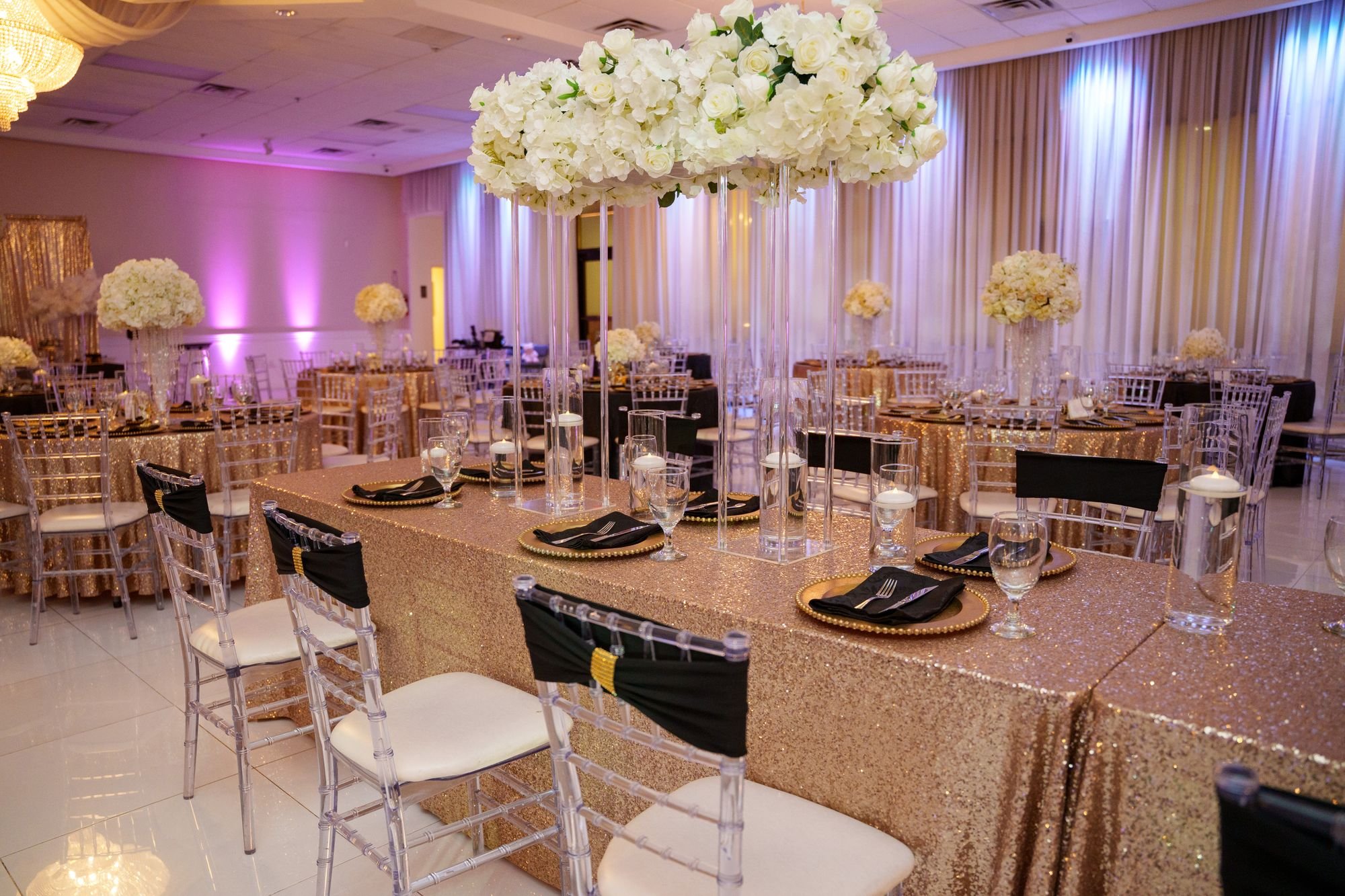 Quinceañera reception ballroom set with rose gold sequin linens black napkins and white rose centerpieces under crystal chandeliers at Luxor Banquet Hall Carrollton