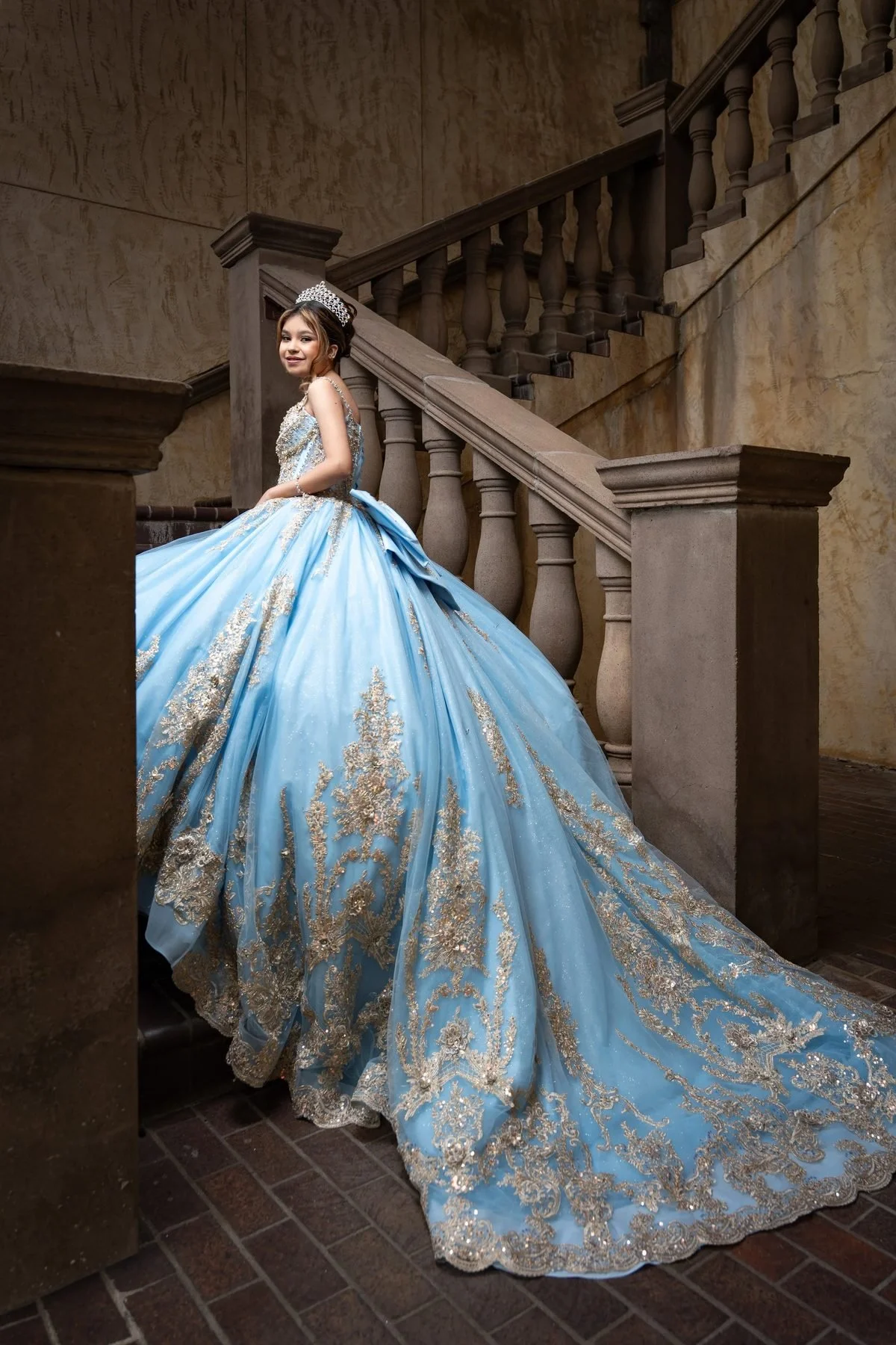 Quinceañera in sky blue gown with gold appliqué and tiara posing on ornate stone staircase