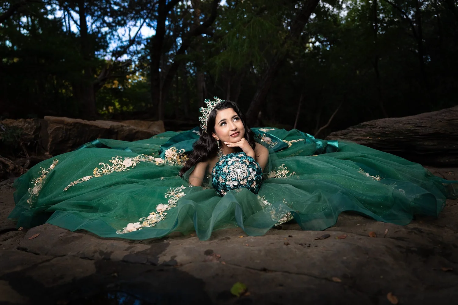 Quinceañera recostada sobre una roca plana del arroyo con ramo de rosas esmeralda, retrato de bosque encantado en Stone Creek Park, Flower Mound, TX