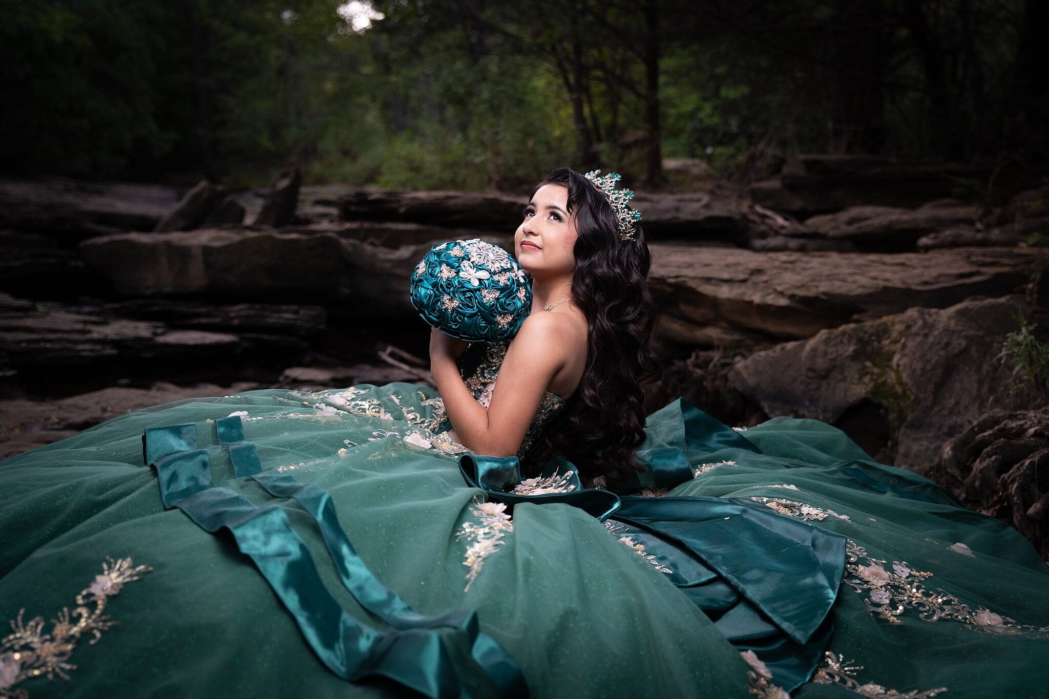  Quinceañera holding emerald rose brooch bouquet and gazing skyward beside layered rock formations at Stone Creek Park in Flower Mound, TX 