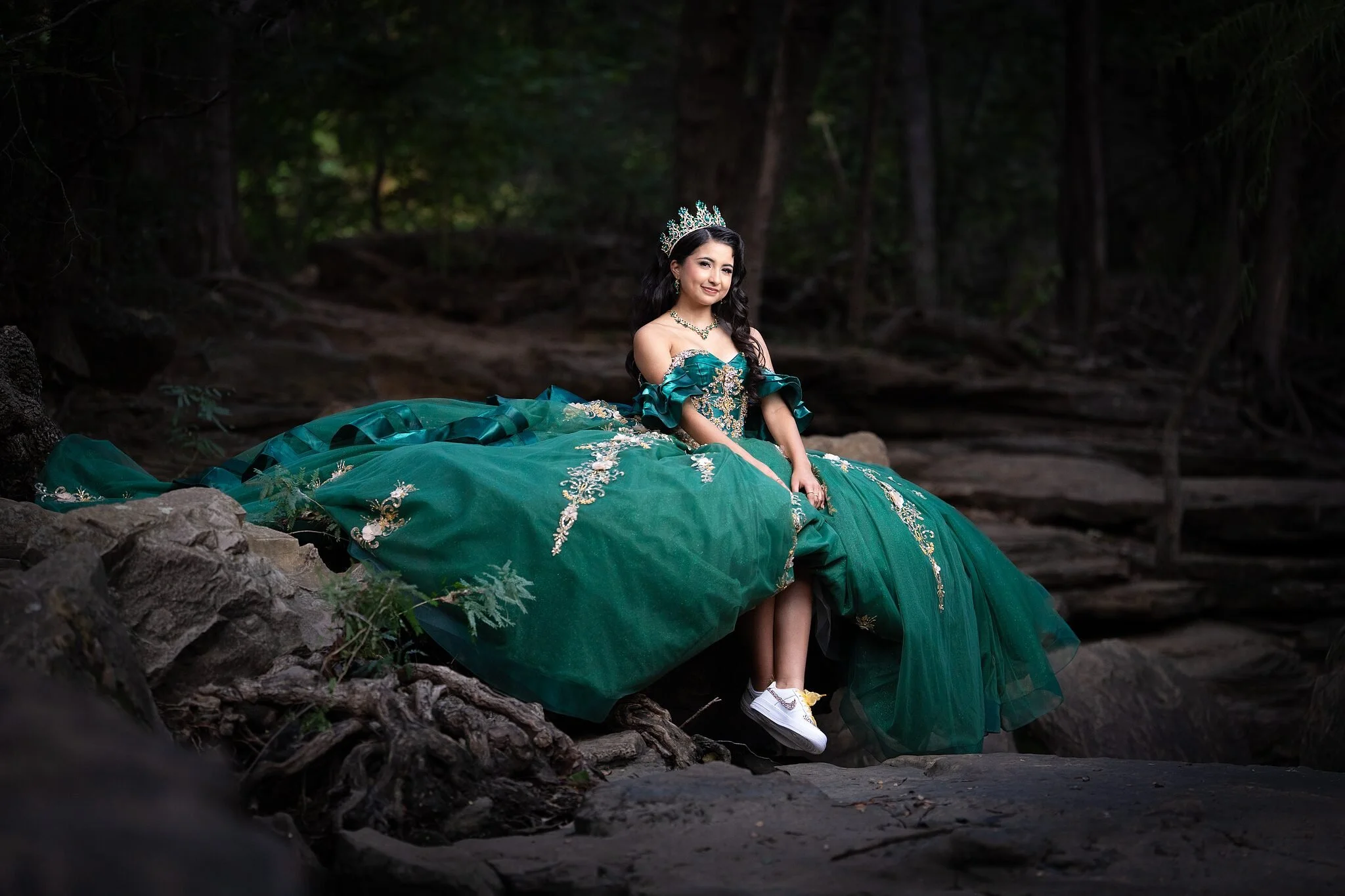  Quinceañera in emerald and gold ball gown sitting on creek rocks with white sneakers peeking from beneath the skirt at Stone Creek Park in Flower Mound, TX 
