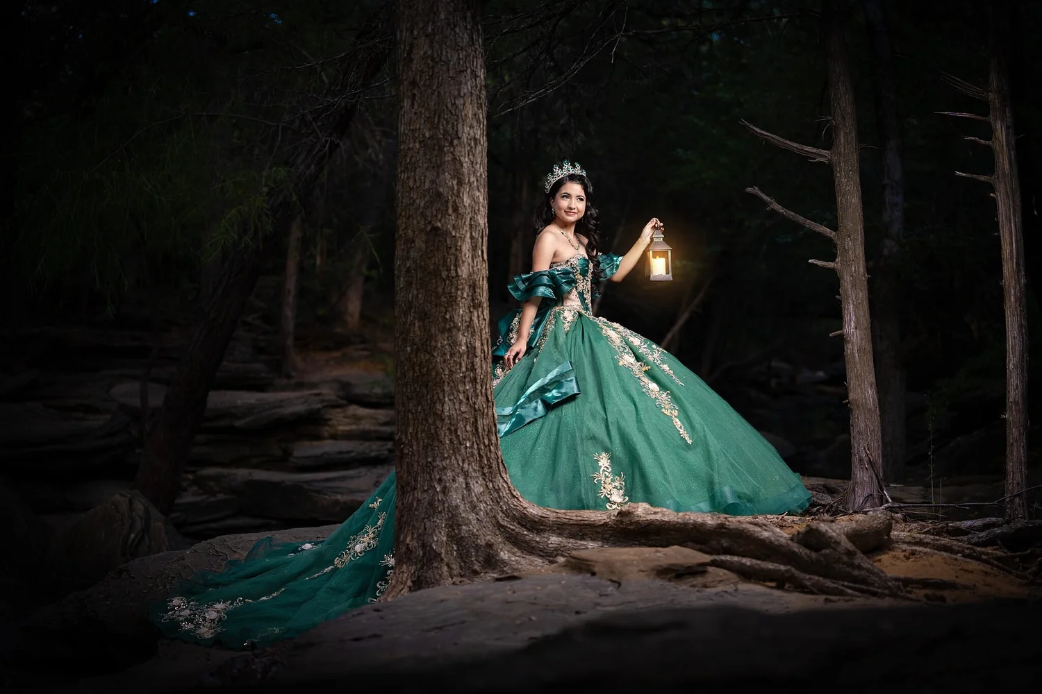 Quinceañera con vestido verde esmeralda y corona de cristal sosteniendo una linterna encendida entre los árboles de Stone Creek Park, tema de bosque encantado en Flower Mound, TX