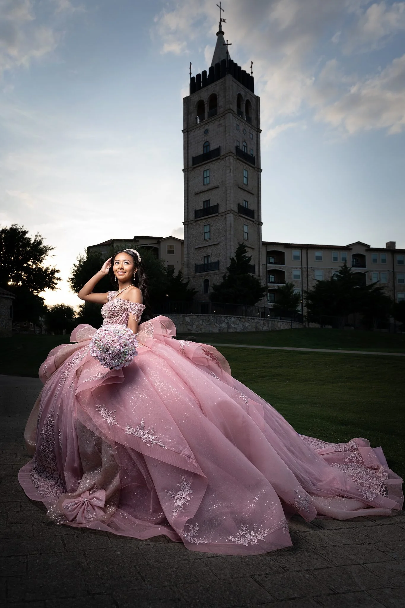 Quinceañera con vestido de gala rosa malva y ramo de joyas, atardecer dorado detrás de la torre del campanario de Adriatica Village en McKinney, TX