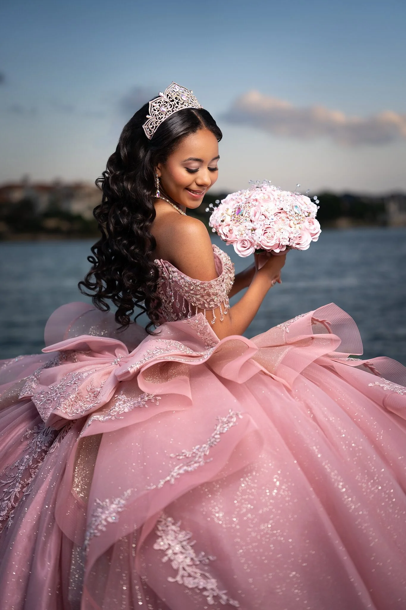  Side profile of quinceañera in pink off-shoulder gown and crystal crown holding a pink rose brooch bouquet by the lake at Adriatica Village, McKinney, TX 