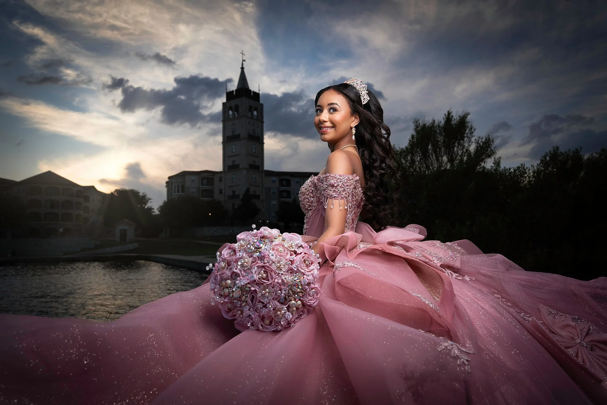  Quinceañera in pink ball gown with sweeping train and floral train detail standing before the Adriatica Village bell tower in McKinney, TX 
