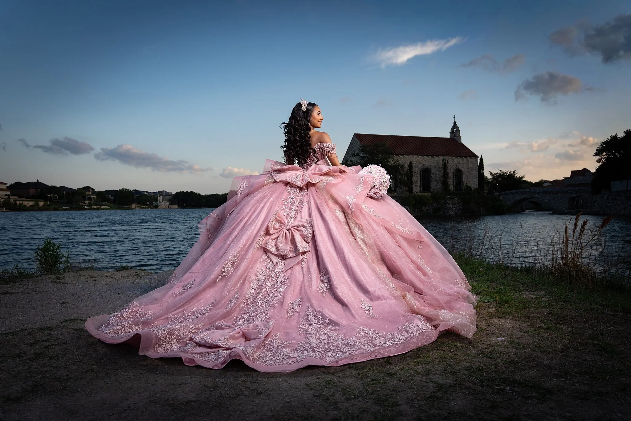  Back view of quinceañera in rose pink ball gown with oversized bow detail, looking across Stonebridge Lake toward Bella Donna Chapel at Adriatica Village, McKinney, TX 
