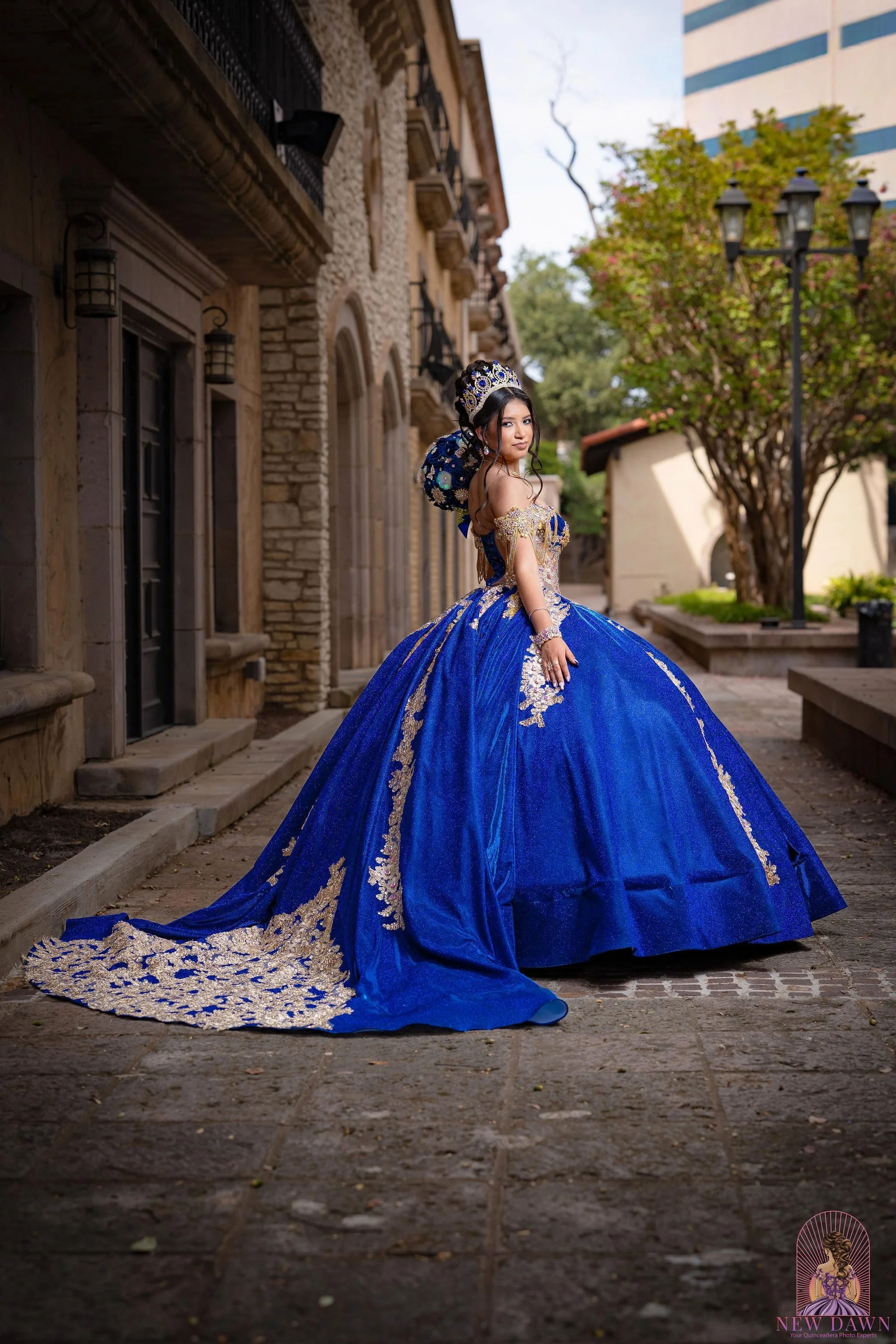 Quinceañera in royal blue and gold ball gown with sweeping train on a cobblestone walkway between Mediterranean buildings at Mandalay Canal Walk in Las Colinas 