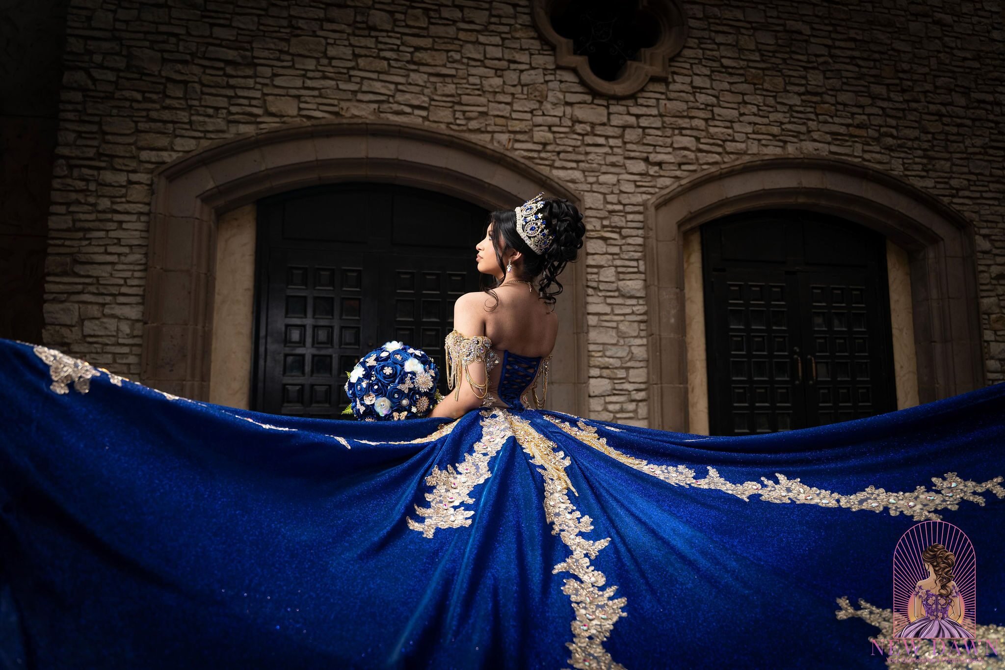  Quinceañera with royal blue and gold ball gown train spread before arched stone doors and limestone wall at Las Colinas / Mandalay Canal 