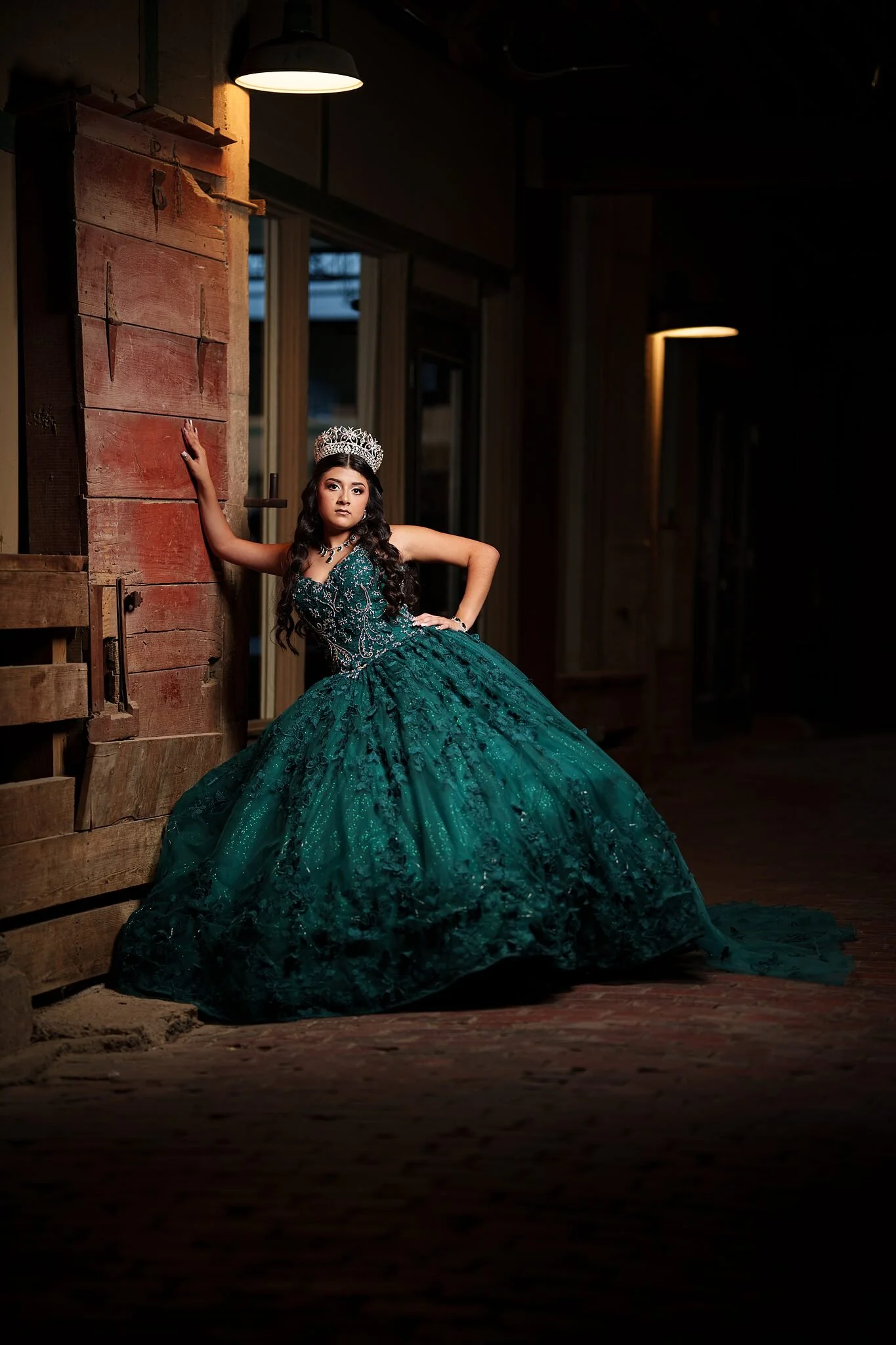  Quinceañera in emerald ball gown leaning against a weathered red wooden door beneath a vintage hanging barn lamp at the Fort Worth Stockyards 