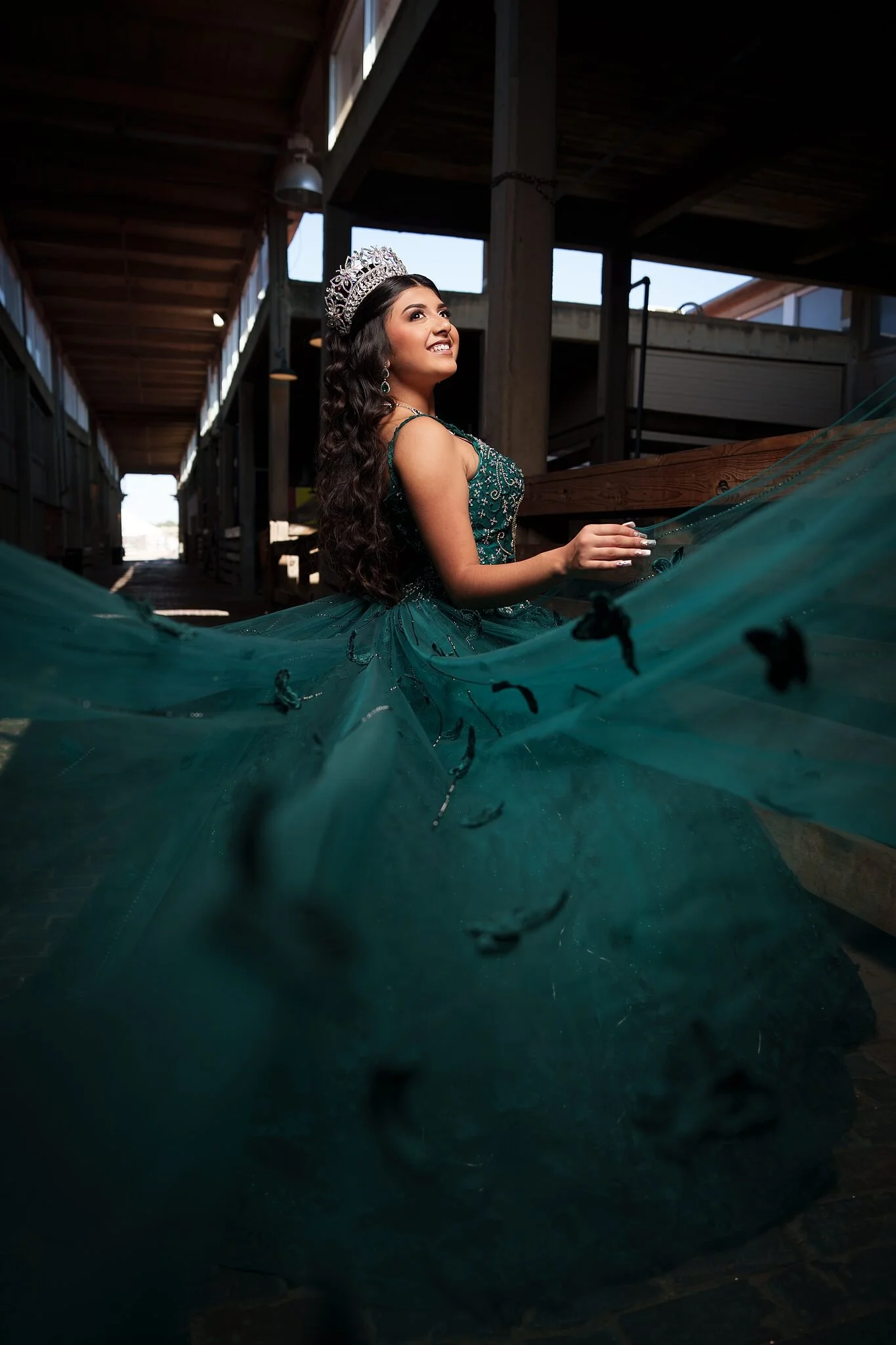  Quinceañera with emerald butterfly-detail train fanned out down a wooden cattle stall corridor at the Fort Worth Stockyards 