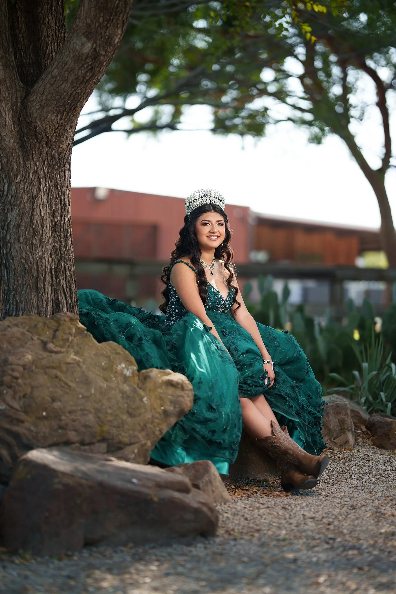 Quinceañera con vestido esmeralda y botas vaqueras sentada en las rocas bajo un árbol en el River Ranch Cactus Garden del Fort Worth Stockyards