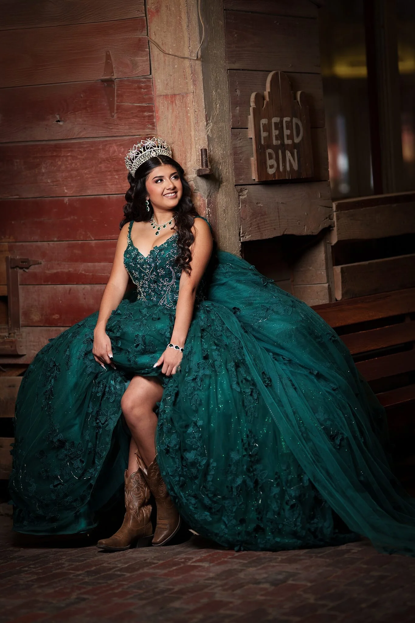  Quinceañera in emerald green ball gown with cowboy boots seated against a weathered red wooden wall beside a vintage "FEED BIN" sign at the Fort Worth Stockyards 