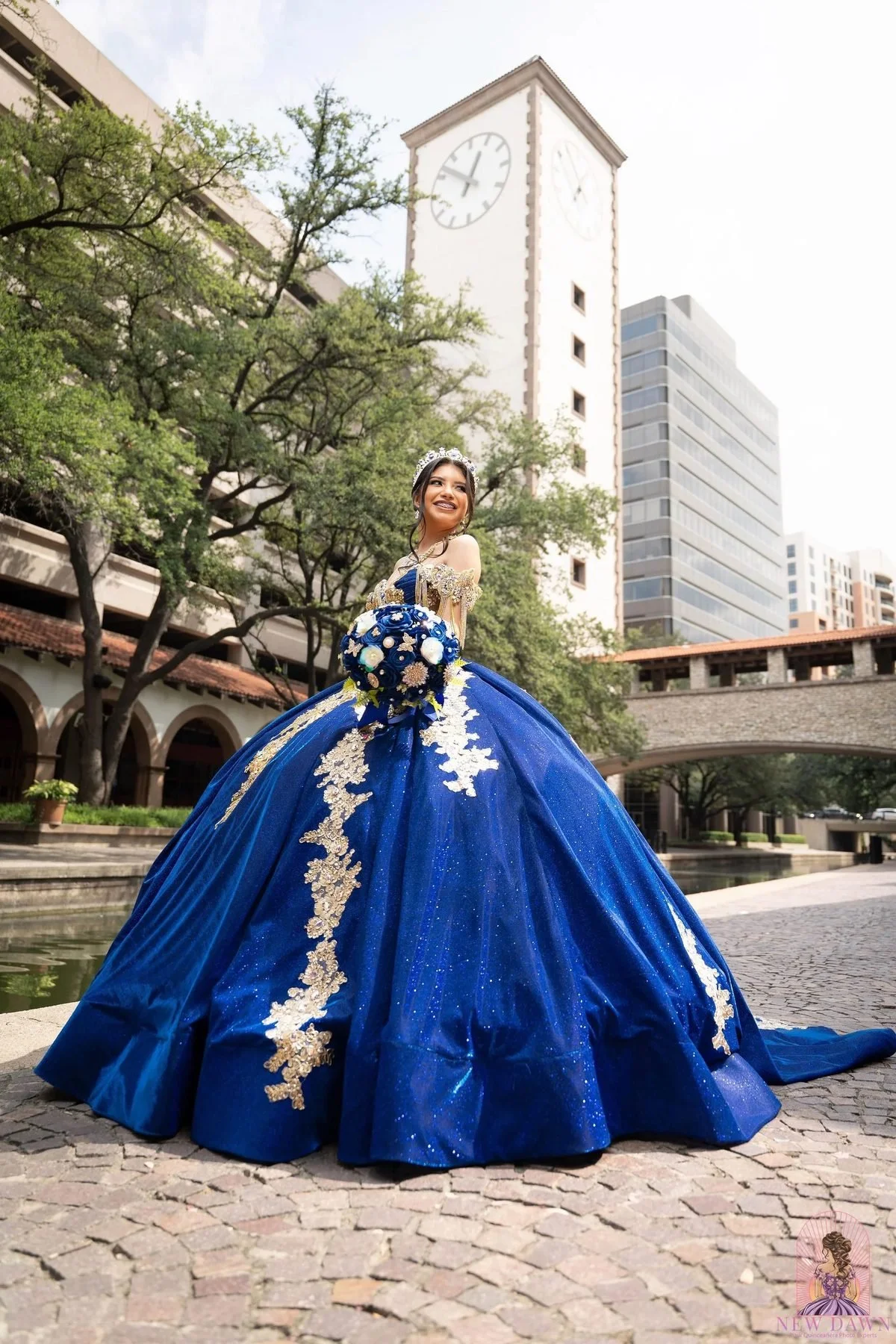 Quinceañera con vestido azul rey y dorado junto a la torre del reloj del Mandalay Canal en Las Colinas, Irving TX