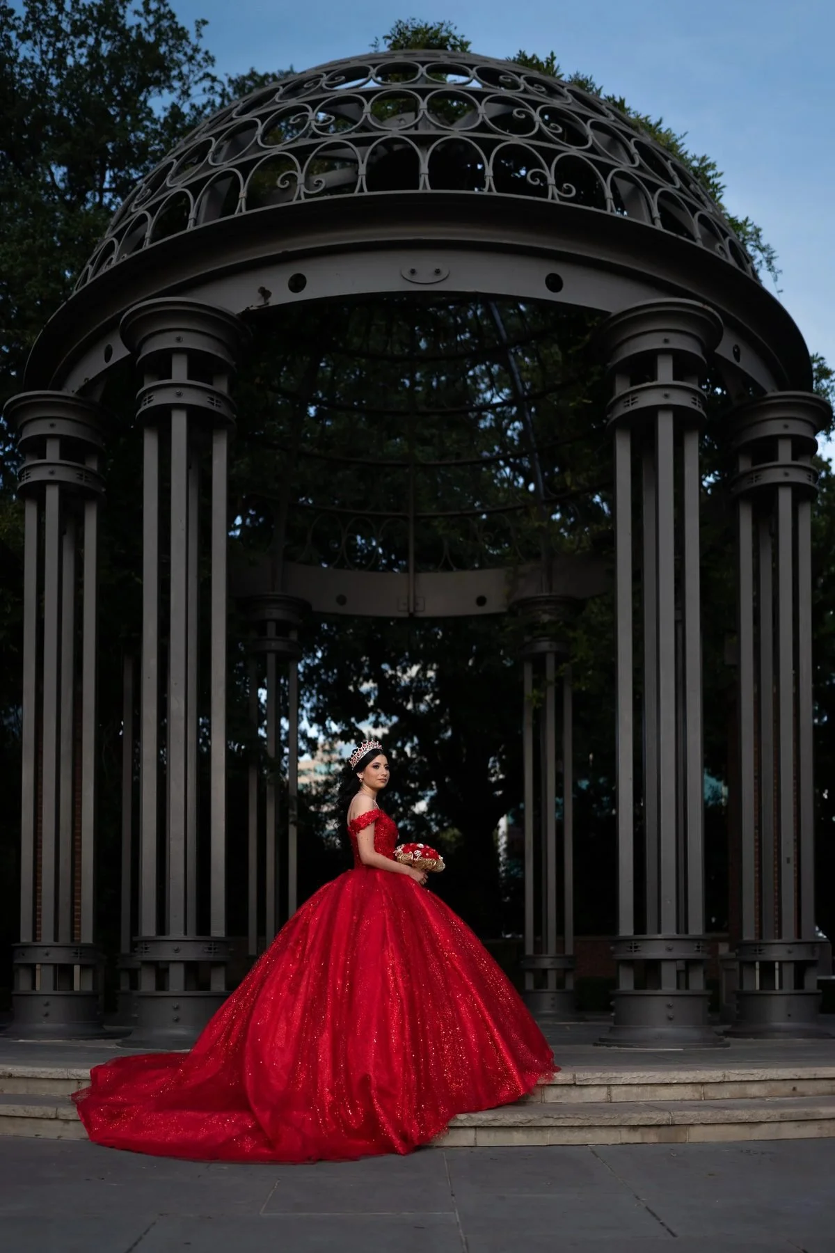 Quinceañera in red sequin gown holding bouquet beneath a domed metal gazebo at a DFW park