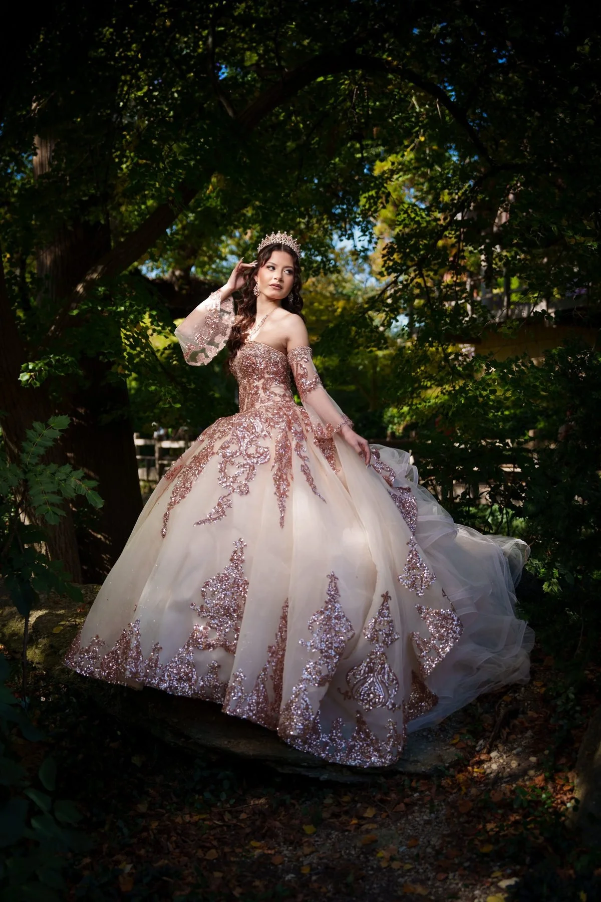 Quinceañera in rose gold sequined ball gown and crown standing under tree archway in Dallas garden