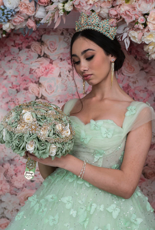Quinceañera wearing a mint green gown and crown, holding an ornate bouquet with pearls and gold details against a pink floral backdrop