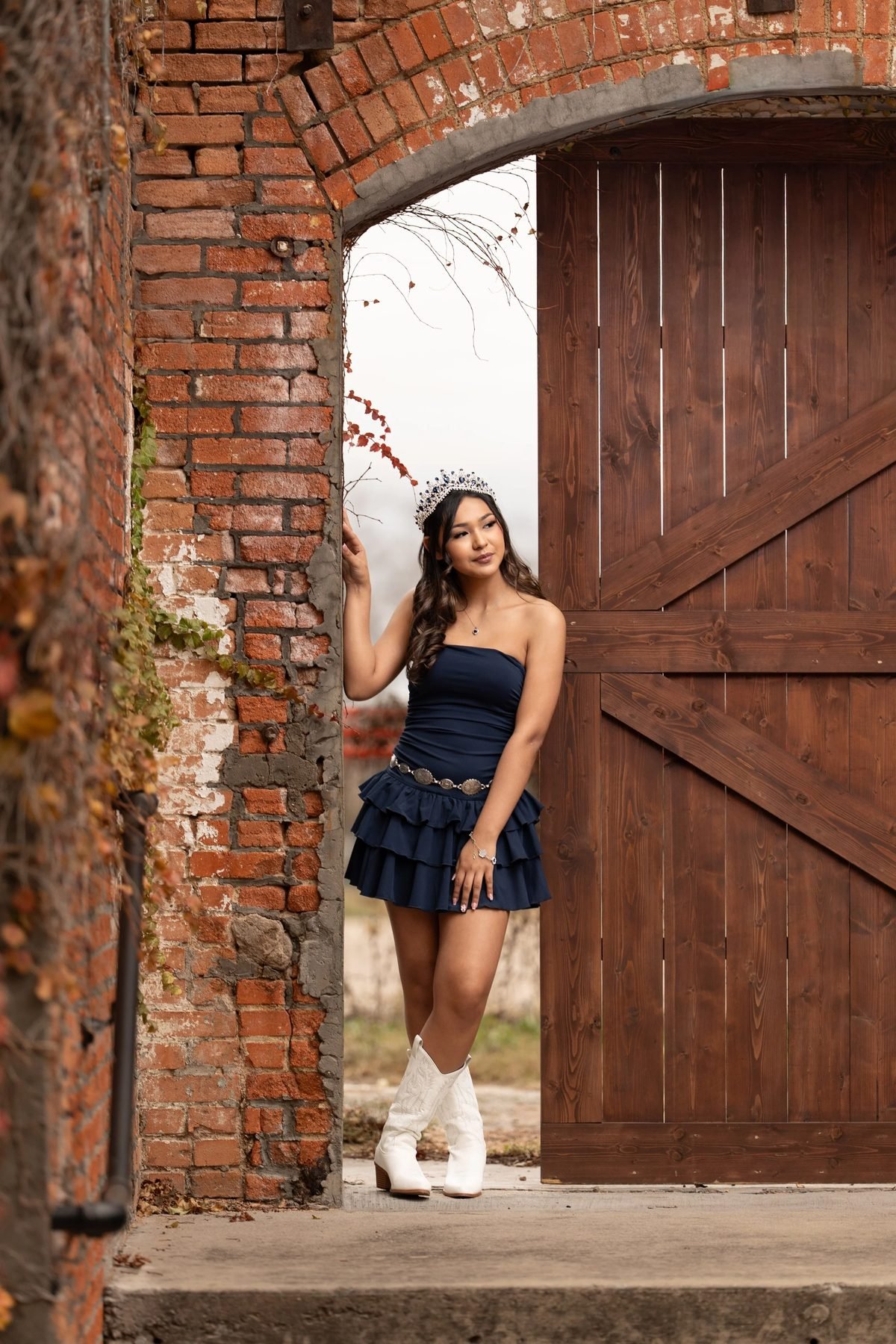 Quinceañera in a navy western romper and white cowboy boots posing in a brick archway for Texas save-the-date photos.