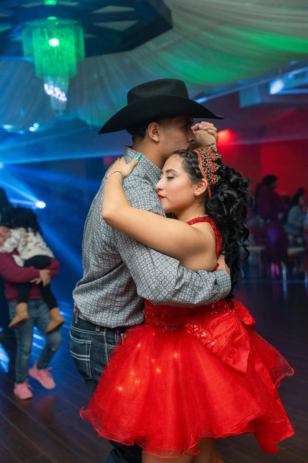 Quinceañera in a red dress dancing with a partner in a cowboy hat under colorful lights at her Texas celebration.