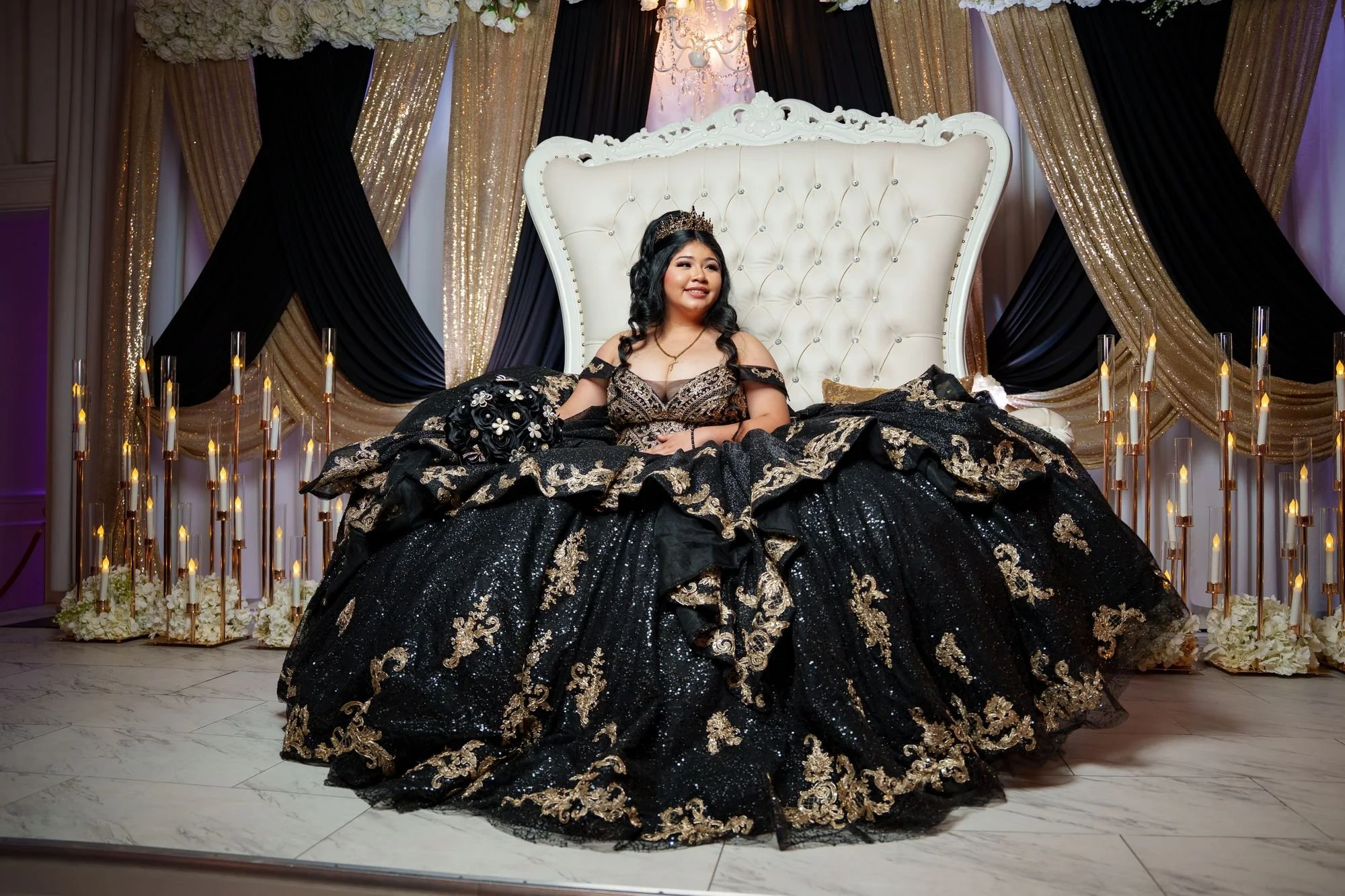 Editorial portrait of Jayden in black and gold quinceañera ballgown seated on white throne with glowing candle tapers and gold marquee 15 in background at Luxor Banquet Hall Carrollton