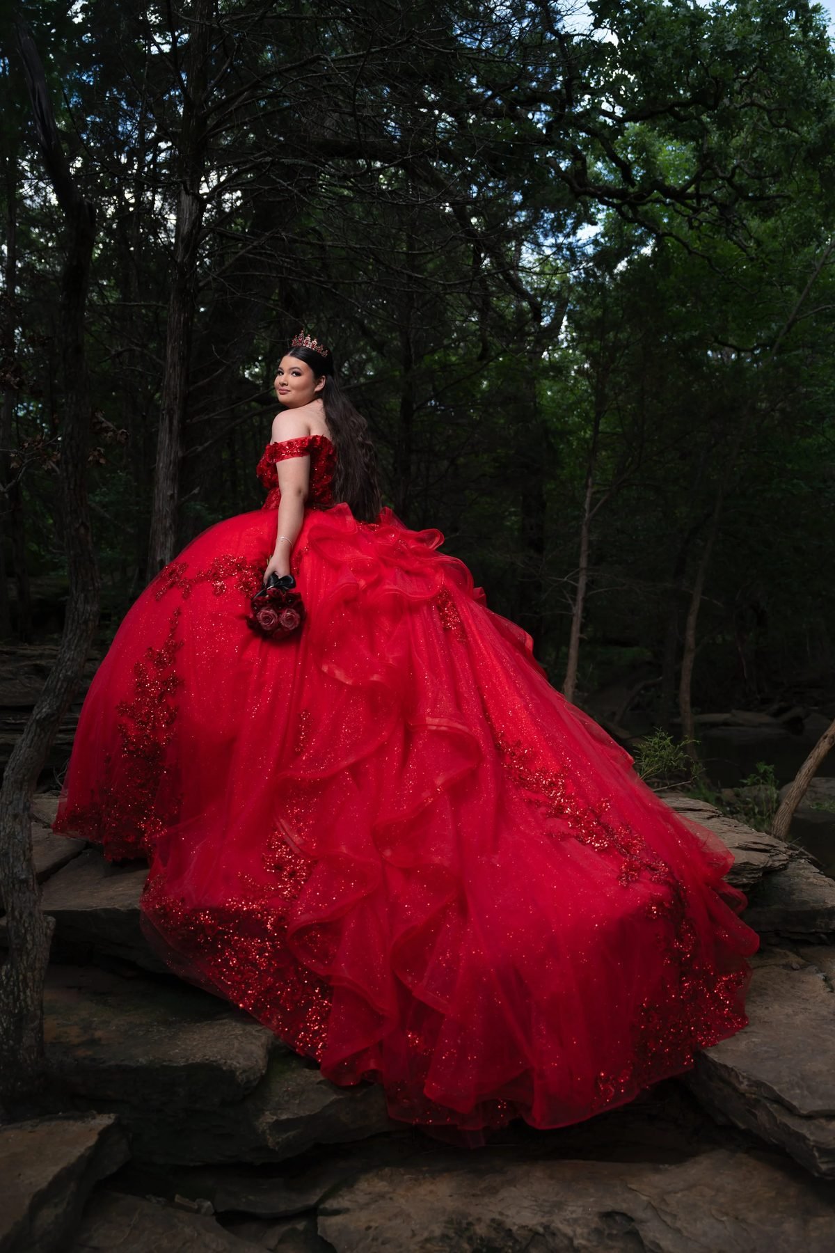 Quinceañera in red ruffled tulle gown holding rose bouquet on forest rocks in DFW