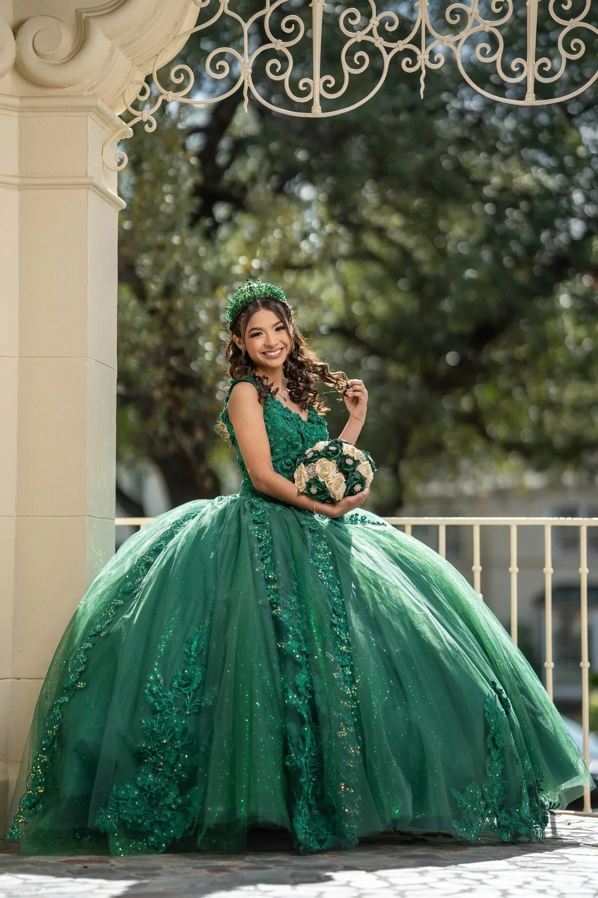 Quinceañera in an emerald green ball gown and crown holding ivory roses in a garden gazebo near Dallas.
