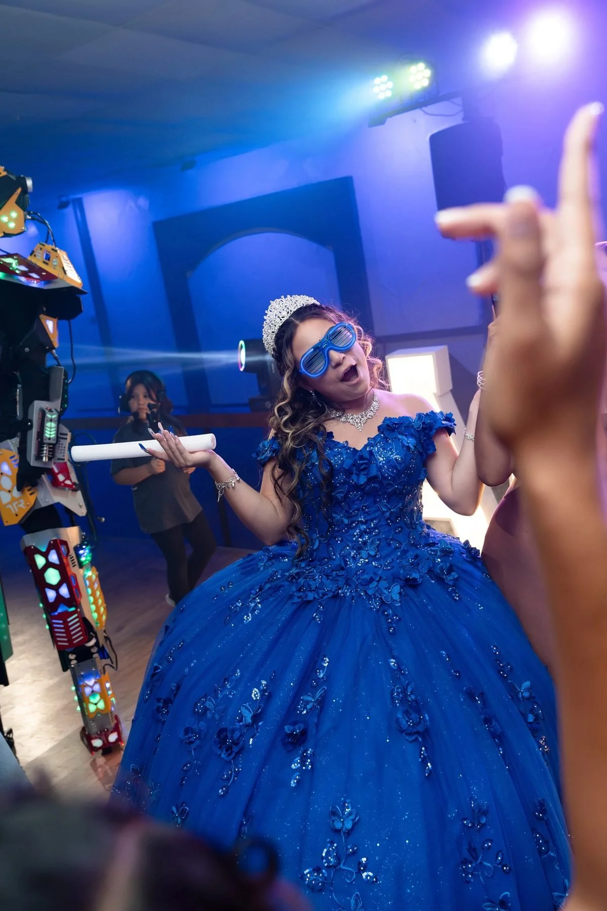 Quinceañera in a royal blue gown and sunglasses dancing with a party robot at her Texas reception.