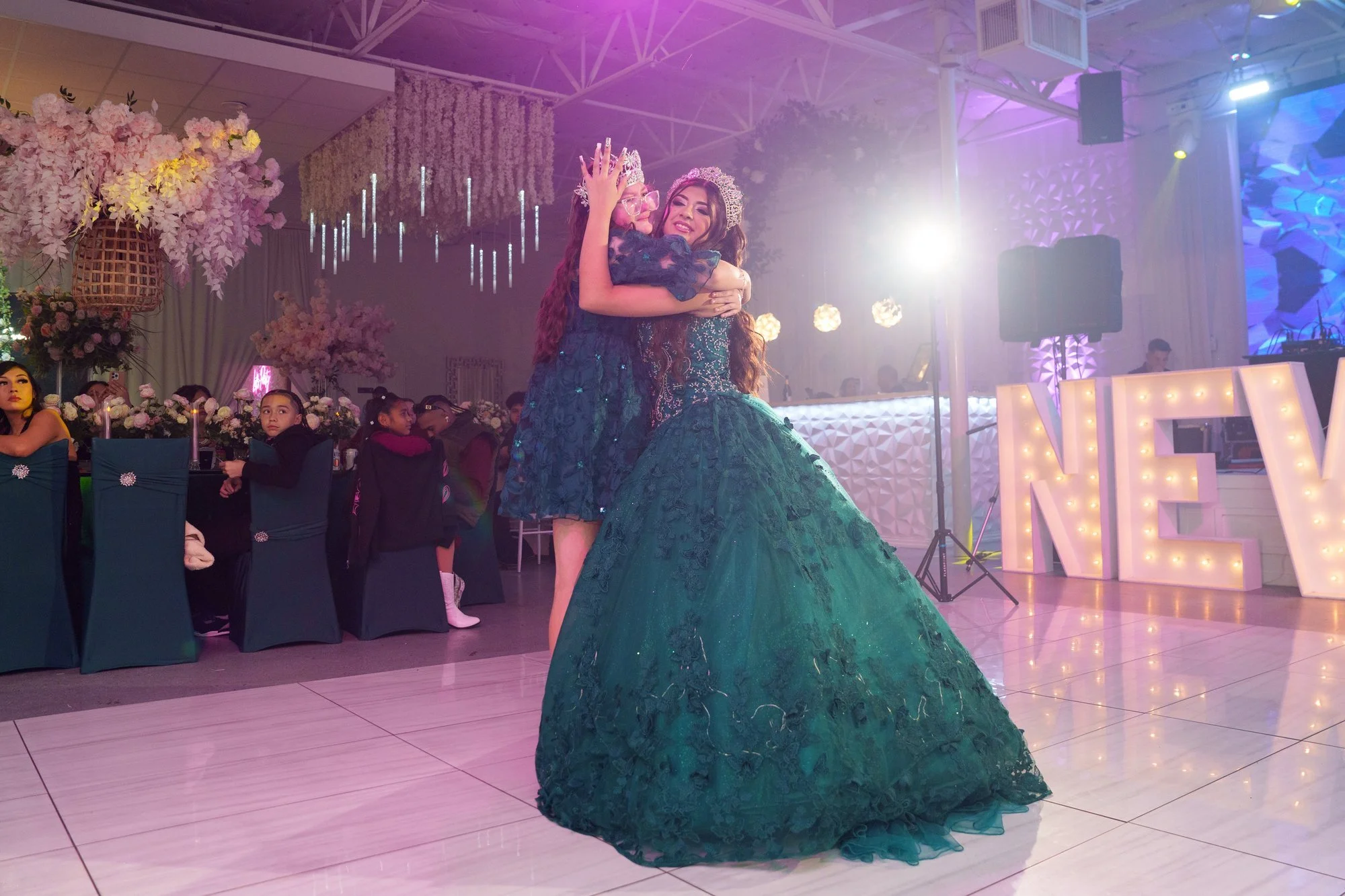 Nevaeh embracing her younger sister moments after the crowning, both in emerald with crowns, beneath a floral and chandelier arch