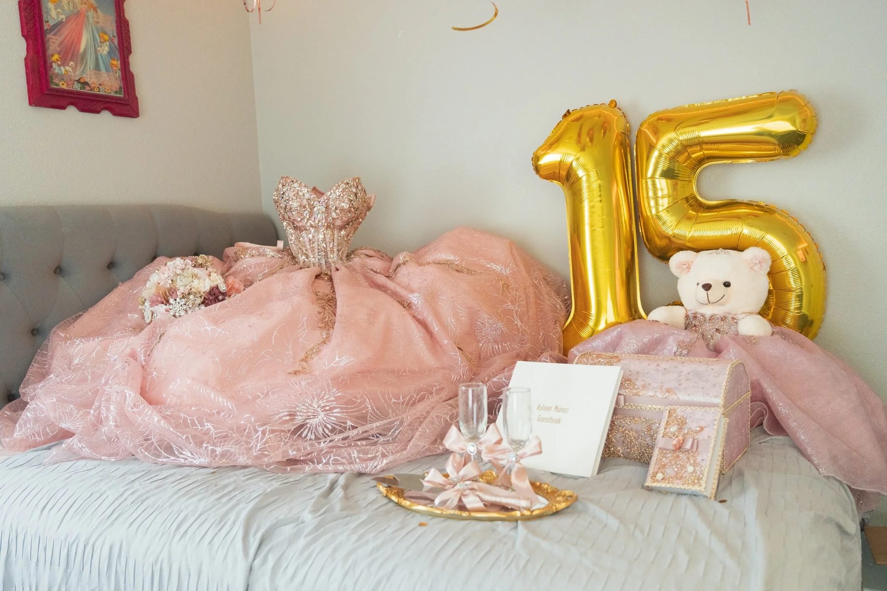 Quinceañera's bedroom décor before the party: blush-pink ball gown, gold "15" balloons, teddy bear, guestbook, and toasting flutes