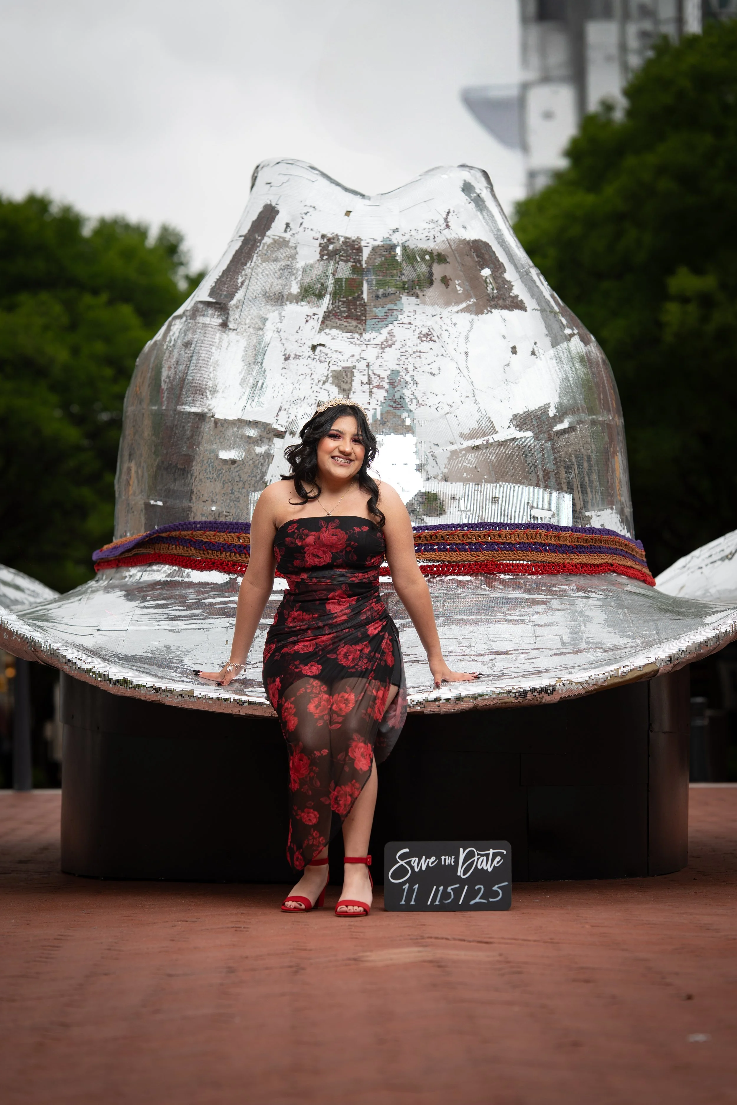 Young woman in a black and red floral dress standing in front of a large shiny metallic sculpture of a hat in an outdoor park setting. She is smiling and posing with a small black sign that has white writing, reading 'Save the Date 11/15/25.'