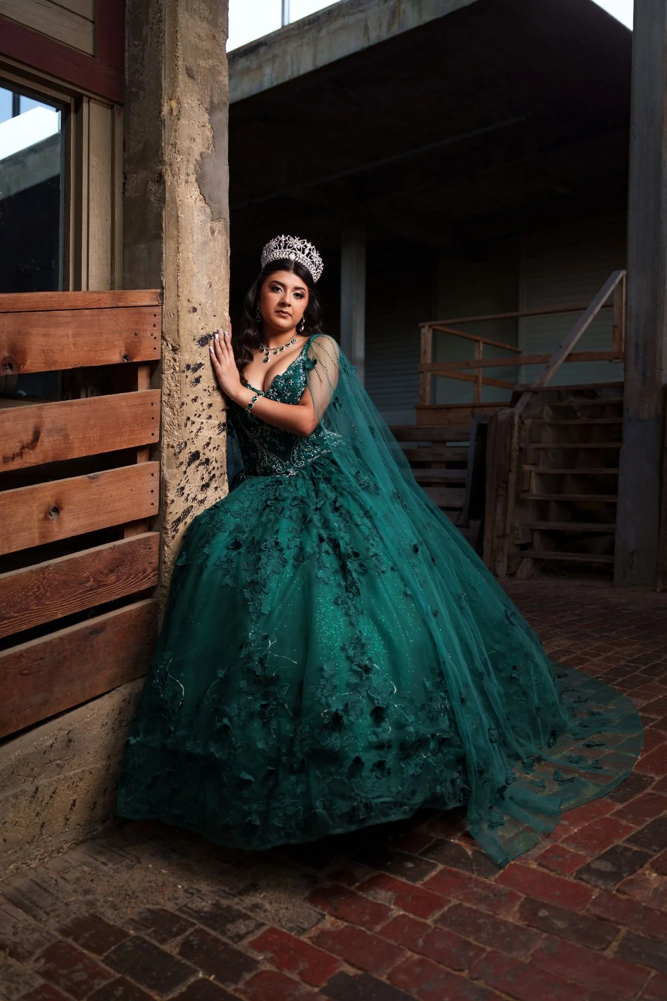 Nevaeh in her emerald quinceañera ballgown with cape and crown, leaning against weathered wood architecture at Fort Worth Stockyards